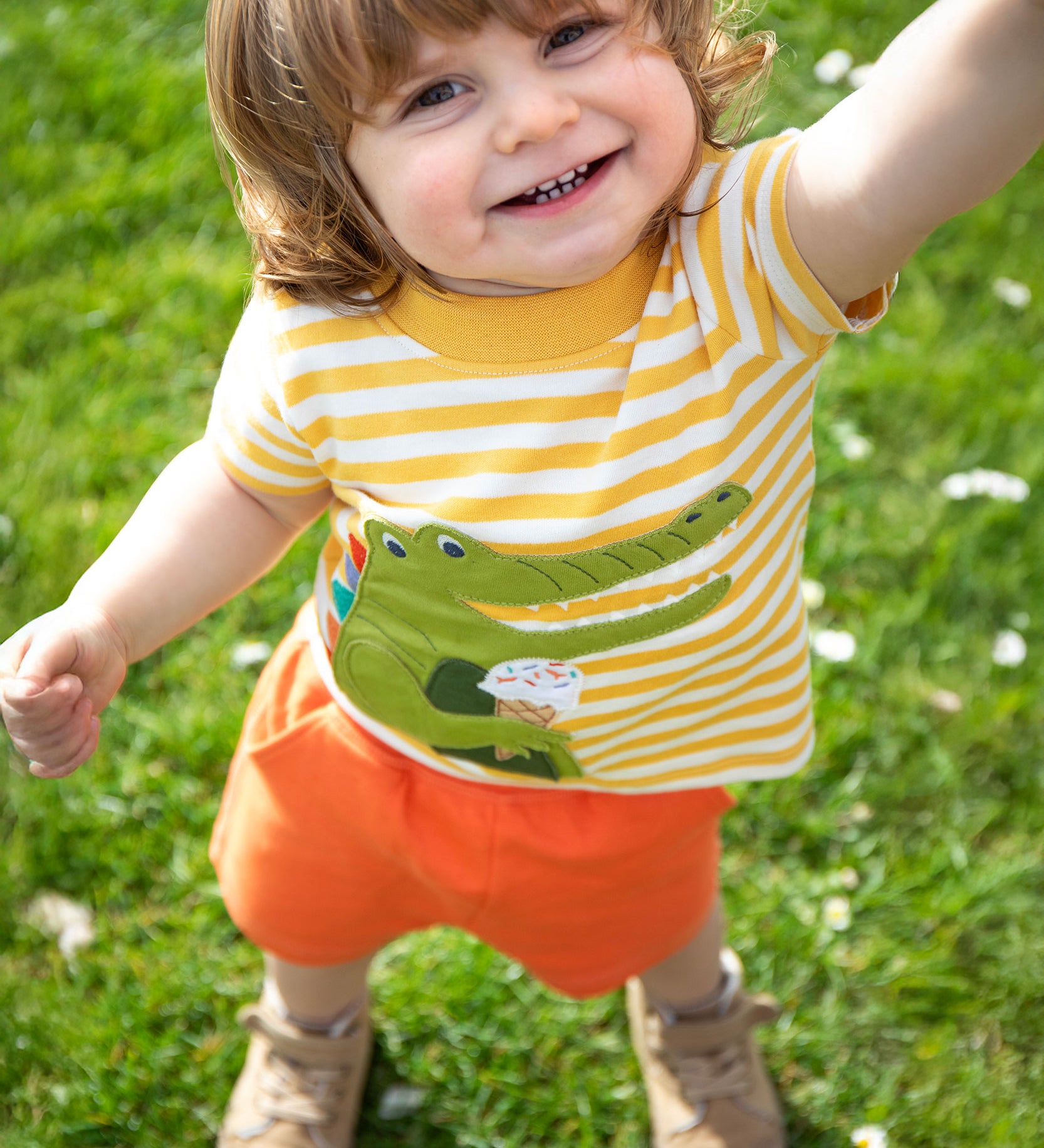 A child wearing the Frugi Crocodile wrap around outfit which includes a yellow striped t-short with a crocodile applique detail and orange coloured shorts.