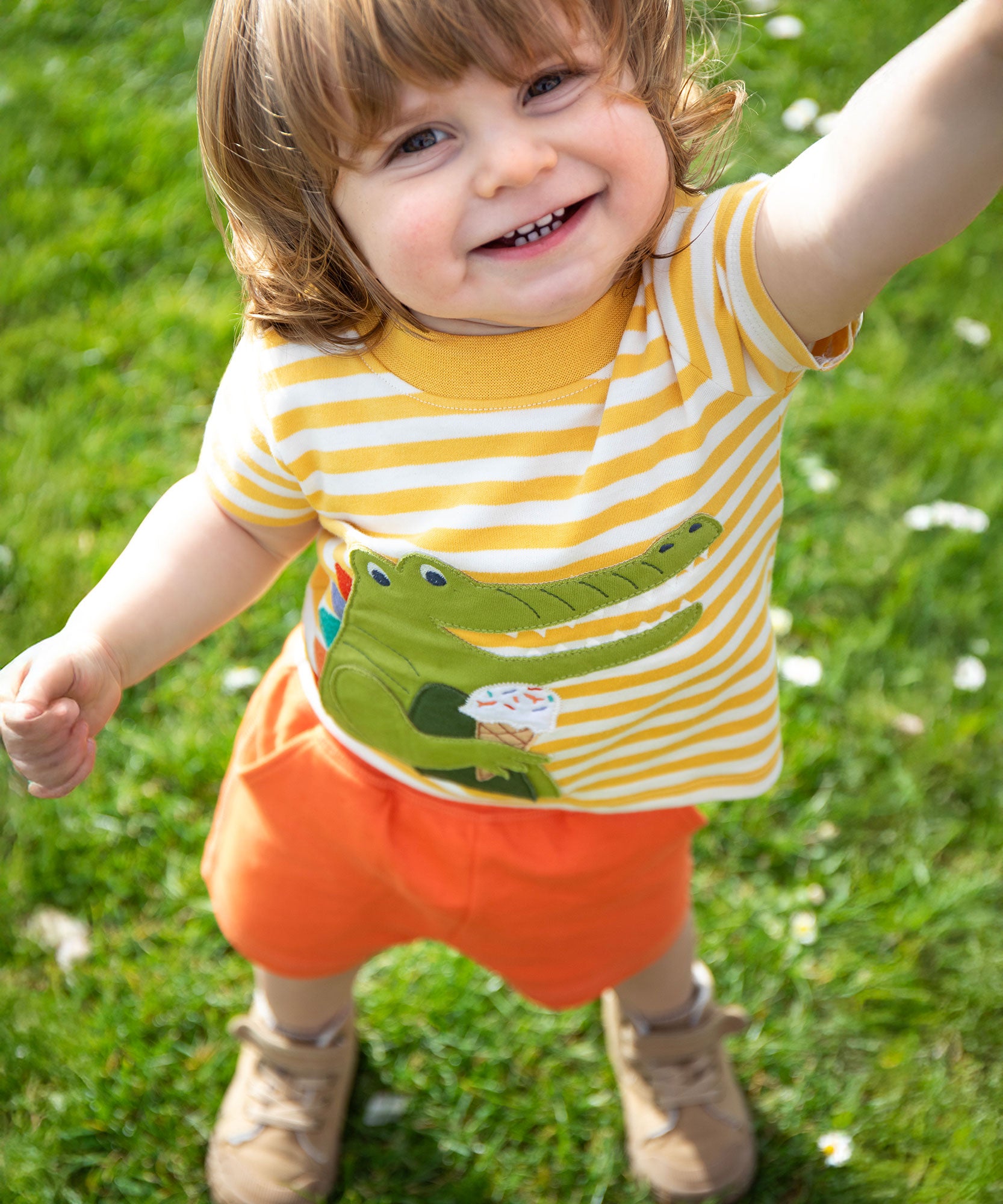 A child wearing the Frugi Crocodile wrap around outfit which includes a yellow striped t-short with a crocodile applique detail and orange coloured shorts.