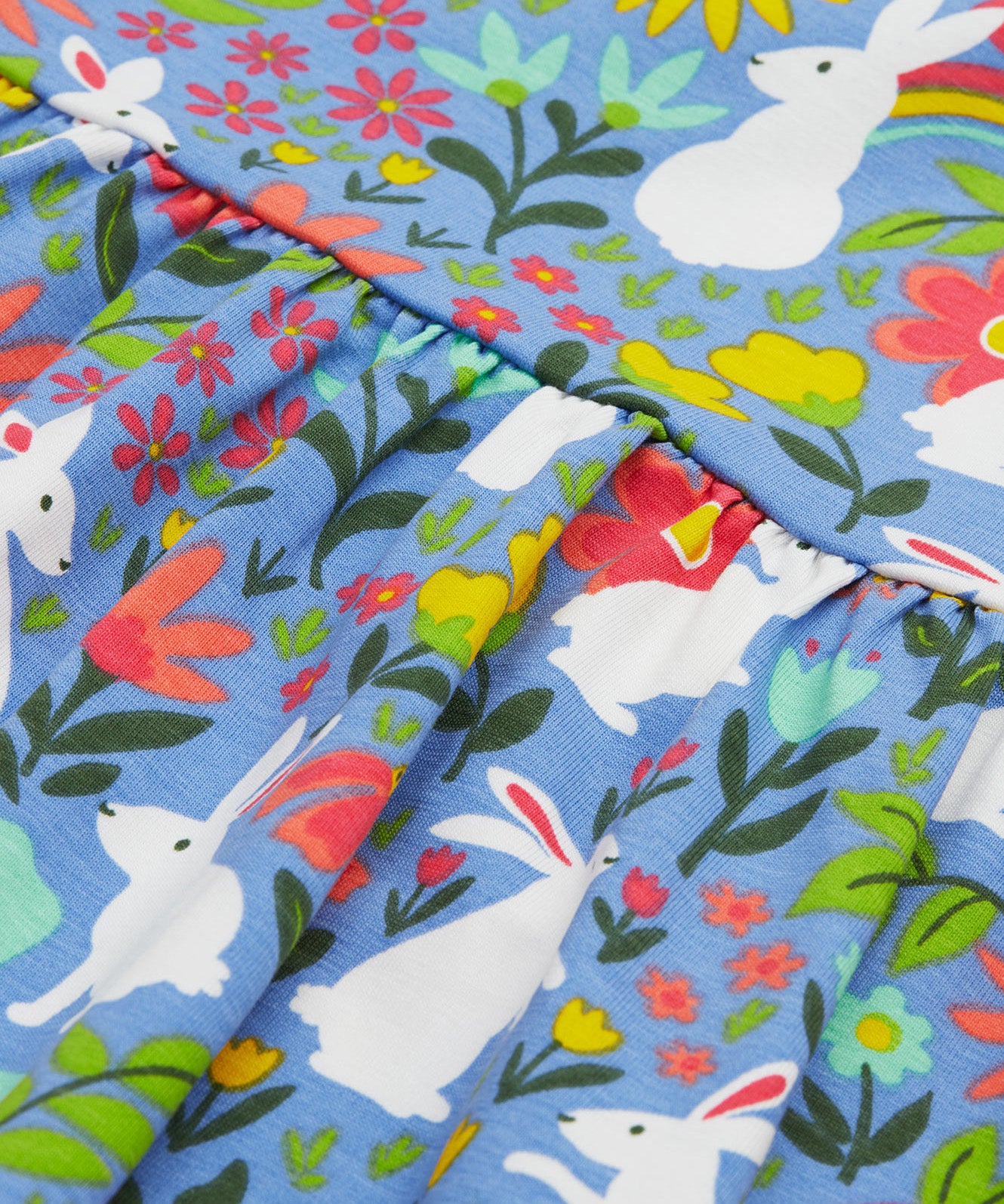 Close up of the Frugi kids Bella dress bunny bounce. Showing a collection of white rabbits printed with different coloured flowers and bright rainbows on light blue organic cotton fabric