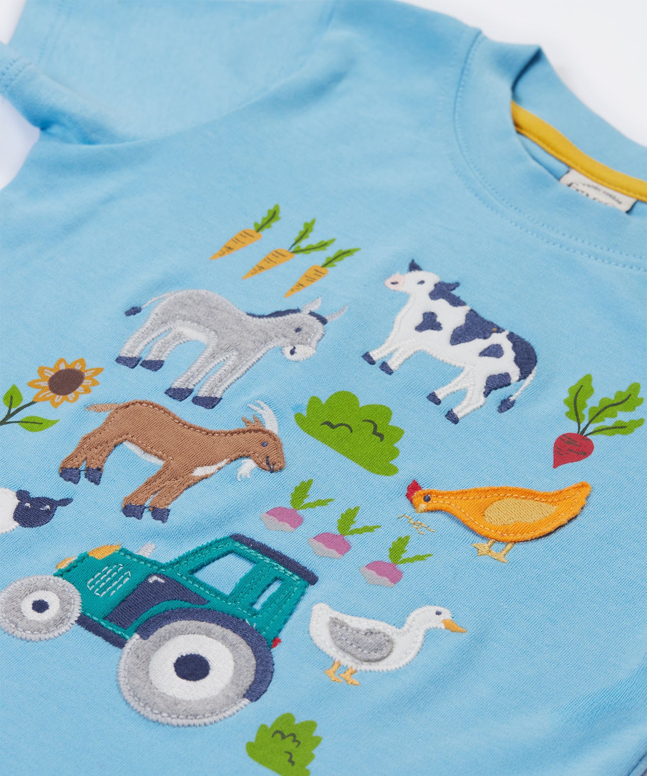 A close up of the Frugi kids bluebird farm life t-shirt  showing the cow, donkey, hen, goat, sheep, goose, tractor, sunflower, carrot, radish and a beetroot appliqué on light blue organic cotton fabric. 