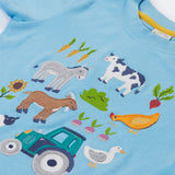 A close up of the Frugi kids bluebird farm life t-shirt  showing the cow, donkey, hen, goat, sheep, goose, tractor, sunflower, carrot, radish and a beetroot appliqué on light blue organic cotton fabric. 