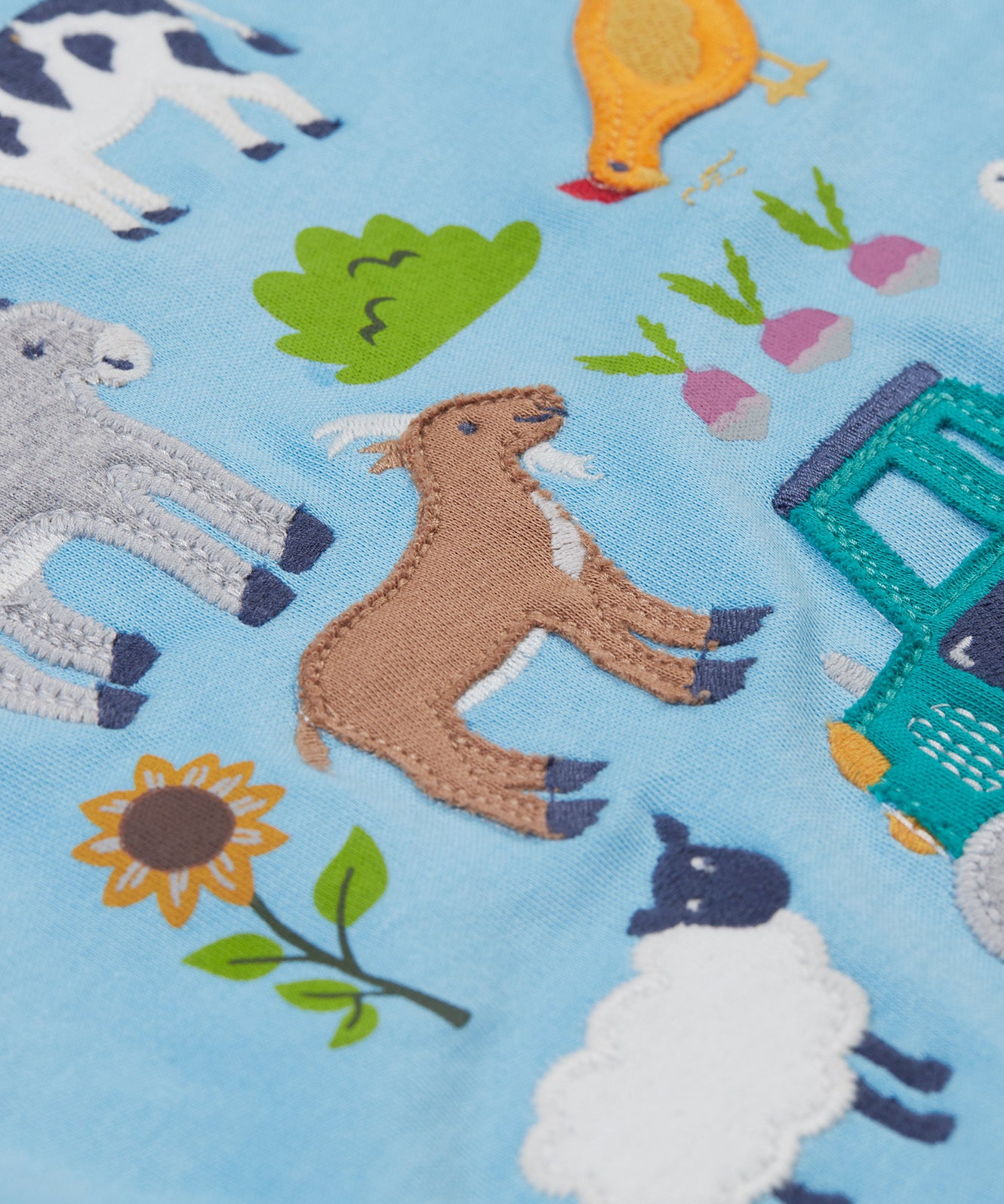 A close up of the Frugi kids bluebird farm life t-shirt  showing the cow, donkey, hen, goat, sheep, goose, tractor, sunflower, carrot, radish and a beetroot appliqué on light blue organic cotton fabric. 