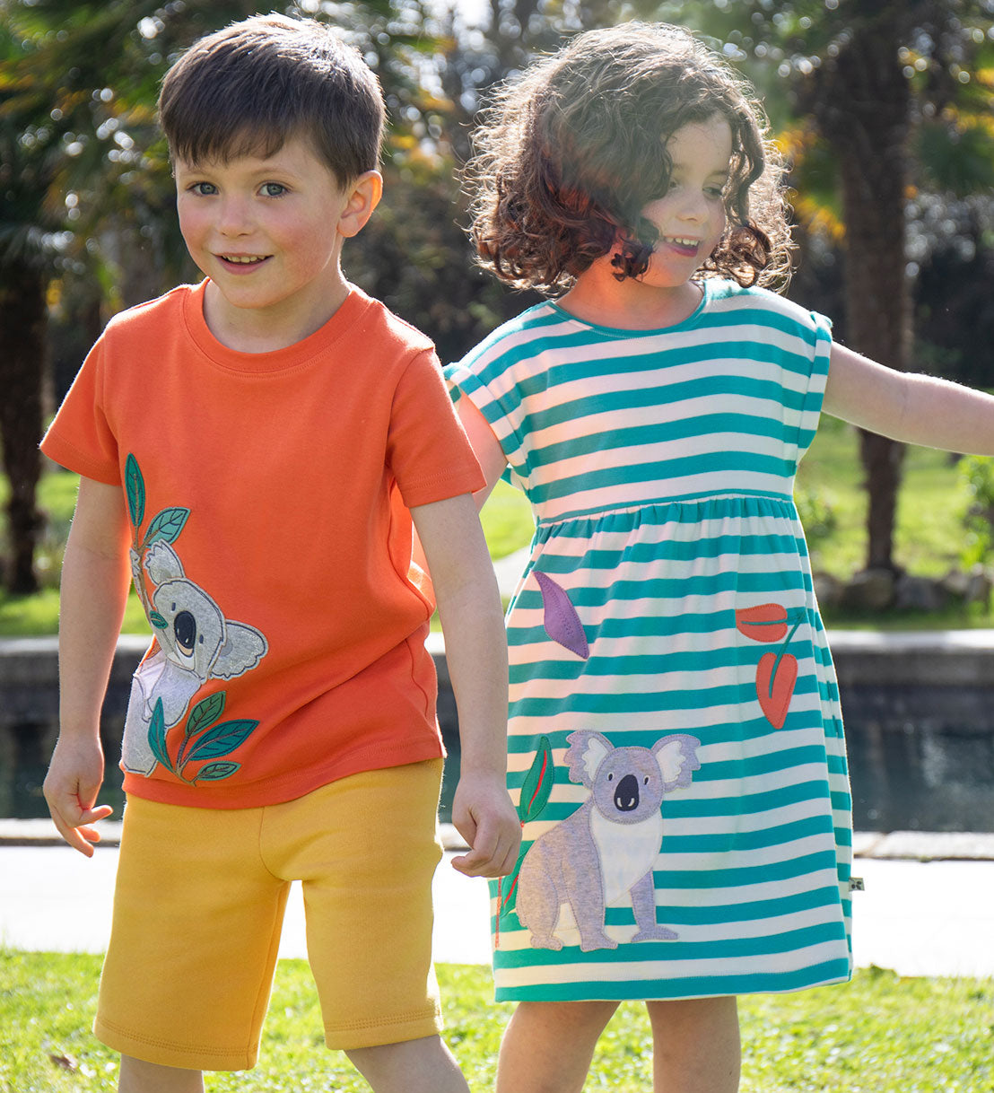 Two children wearing Frugi organic cotton clothing. One is wearing the  Carsen Cool Koala applique t-shirt with yellow bumblebee Samson shorts. The other child is wearing a Koala Felicity Dress.