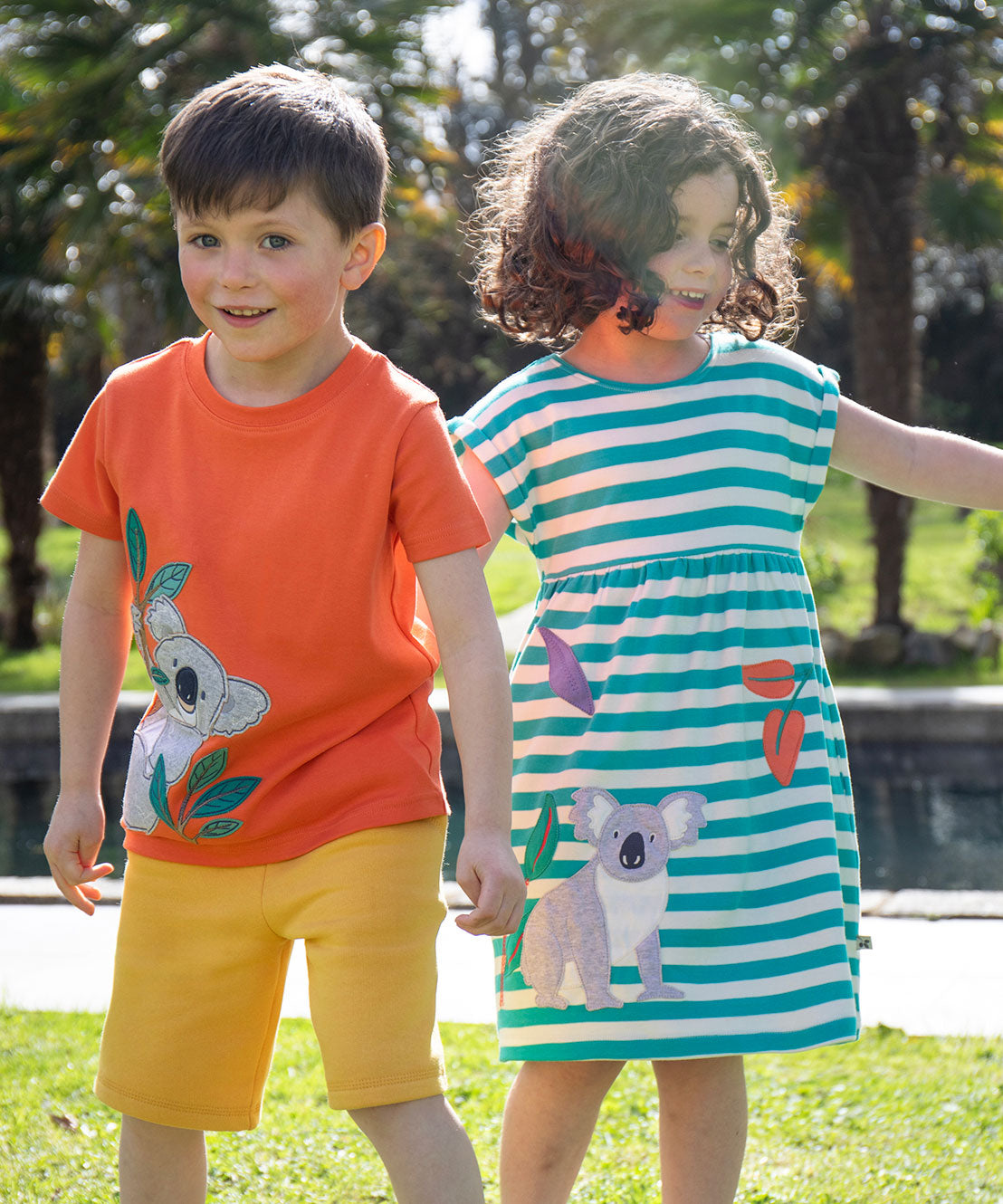 Two children wearing Frugi organic cotton clothing. One is wearing the  Carsen Cool Koala applique t-shirt with yellow bumblebee Samson shorts. The other child is wearing a Koala Felicity Dress.