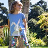 Child leaning against a tree outside wearing the Frugi Cassia t-shirt.  Cornflower blue coloured with a playful appliqué of a grey rabbit with flowers in its mouth with the Frugi Libby Leggings with a floral themed Bunny Bounce print. 