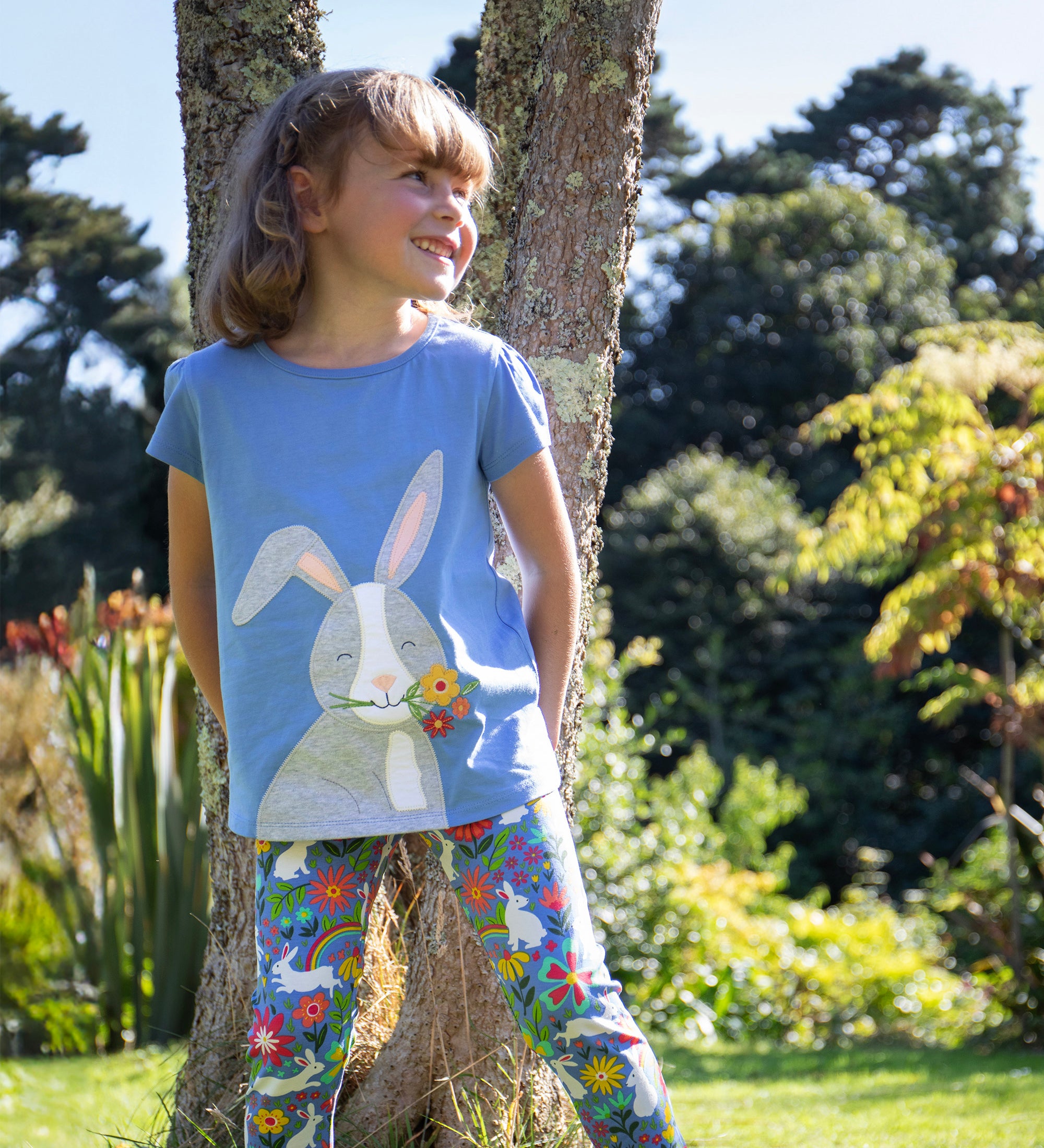 Child leaning against a tree outside wearing the Frugi Cassia t-shirt.  Cornflower blue coloured with a playful appliqué of a grey rabbit with flowers in its mouth with the Frugi Libby Leggings with a floral themed Bunny Bounce print. 