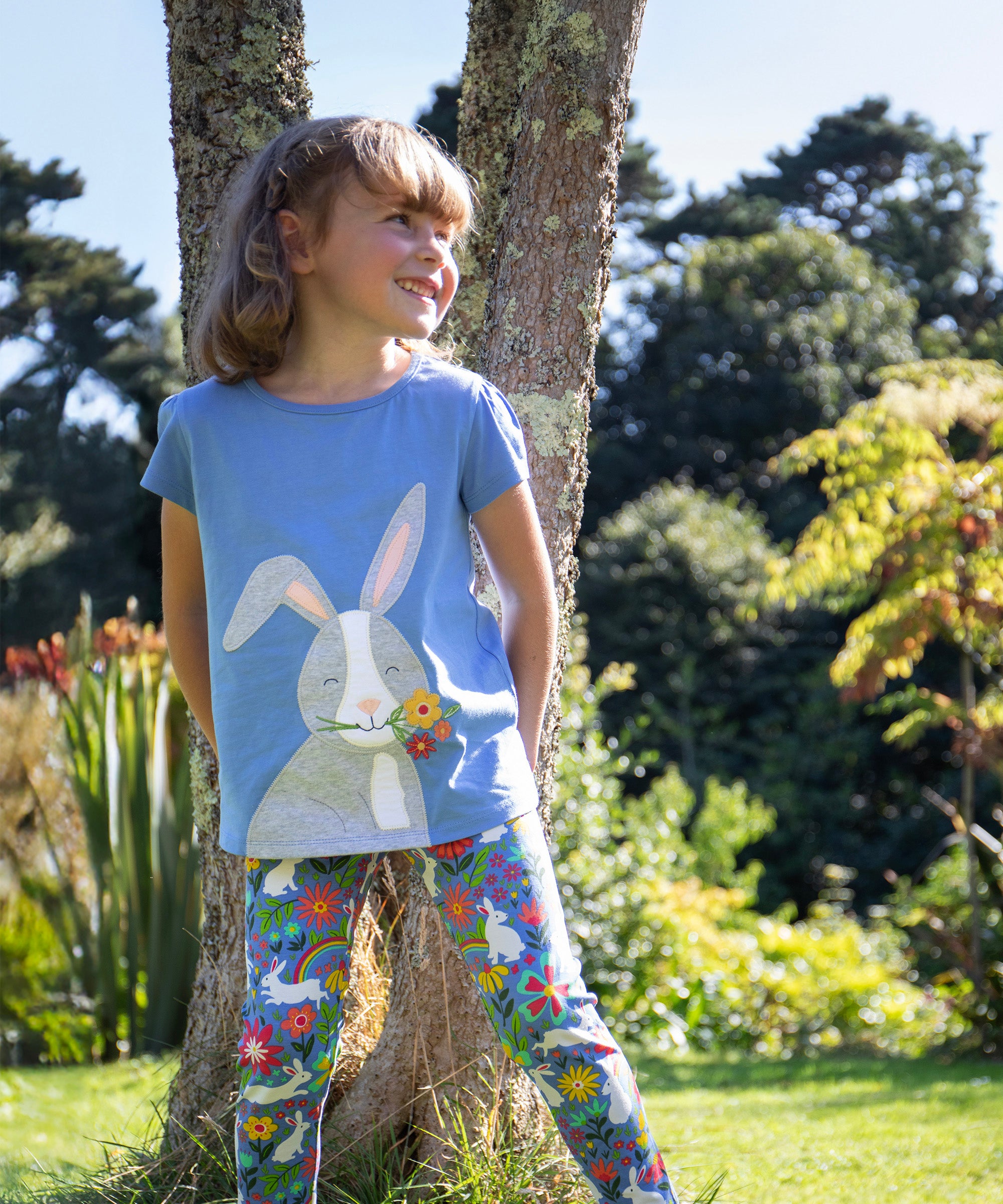 Child leaning against a tree outside wearing the Frugi Cassia t-shirt.  Cornflower blue coloured with a playful appliqué of a grey rabbit with flowers in its mouth with the Frugi Libby Leggings with a floral themed Bunny Bounce print. 