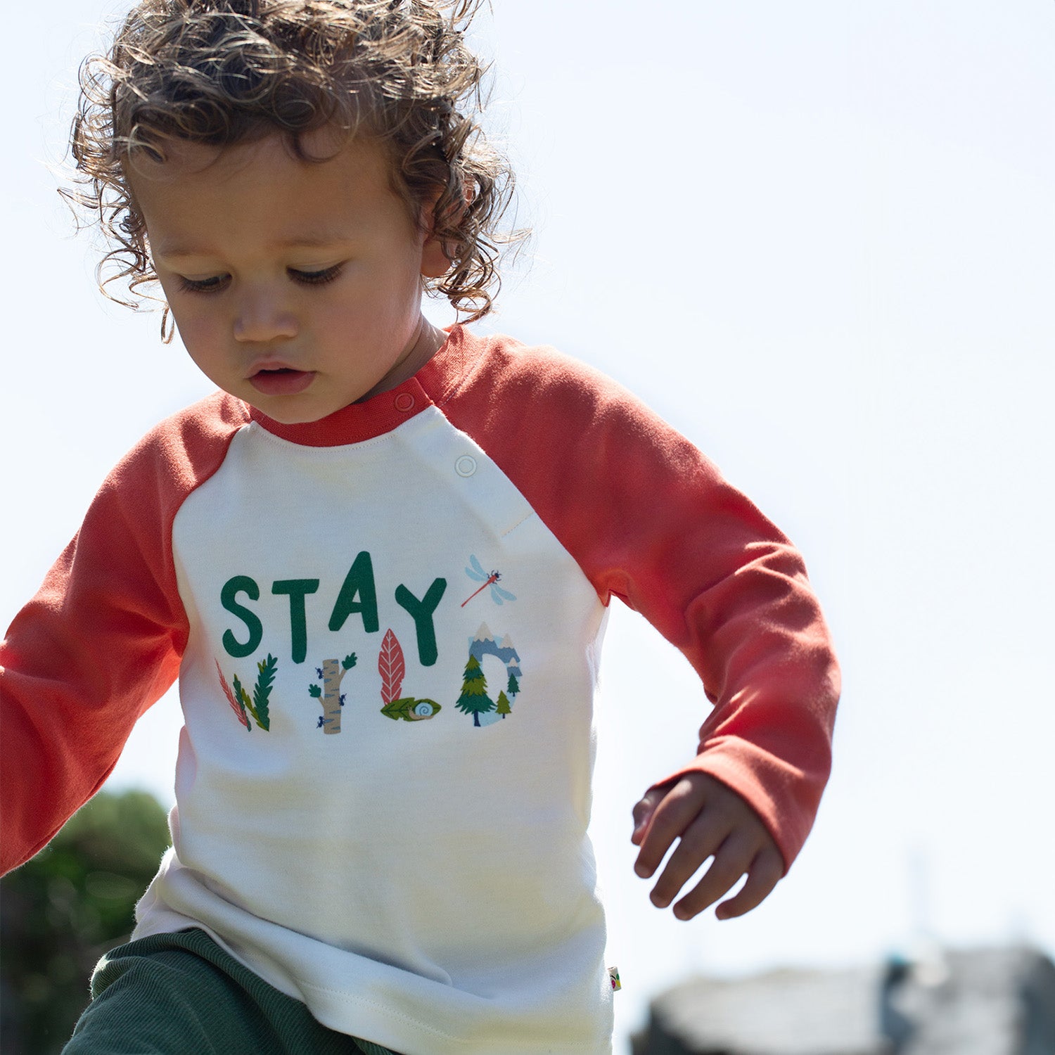 A child wears the Frugi Luke Raglan Top - Stay Wild / Paprika outdoors.