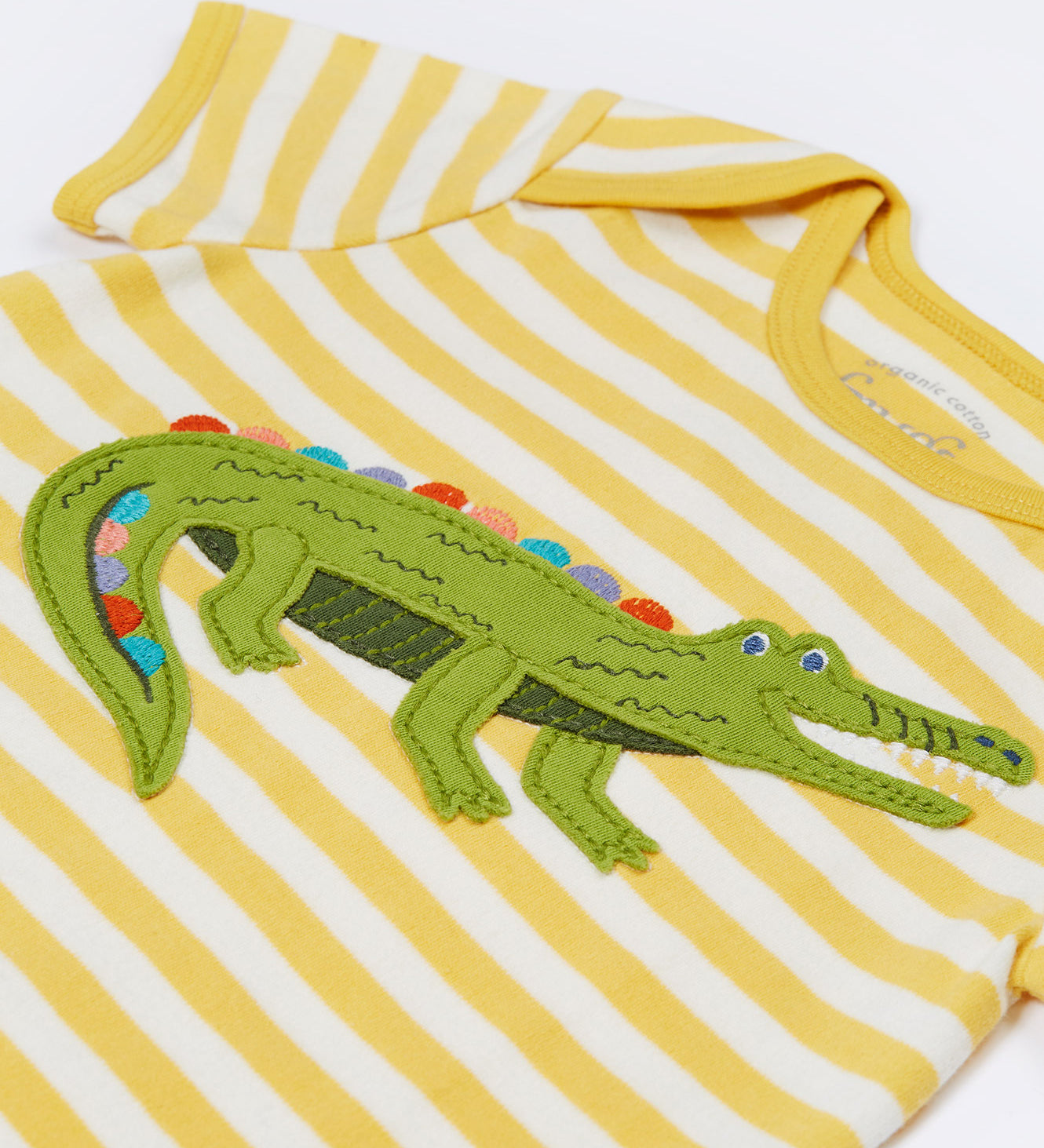 Close up of the Frugi kids crocodile rue romper. Showing the appliqué detail of a green crocodile on yellow and white striped organic cotton fabric