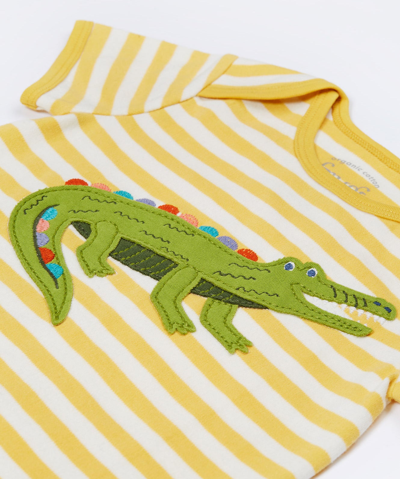 Close up of the Frugi kids crocodile rue romper. Showing the appliqué detail of a green crocodile on yellow and white striped organic cotton fabric