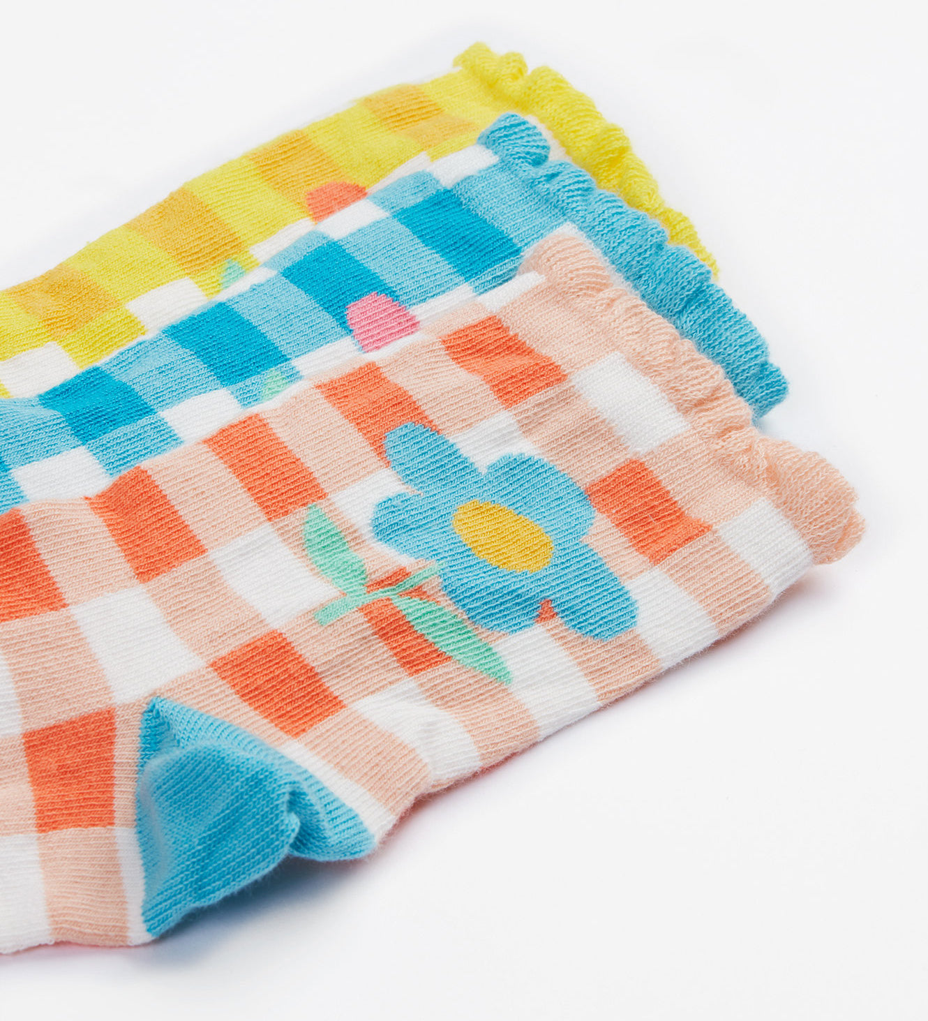 A close up of the Frugi kids Freya frilled gingahm socks - Showing the blue gingham with pink daisy sock, a yellow gingham with light orange daisy sock and a light orange gingham and blue daisy sock on organic cotton fabric