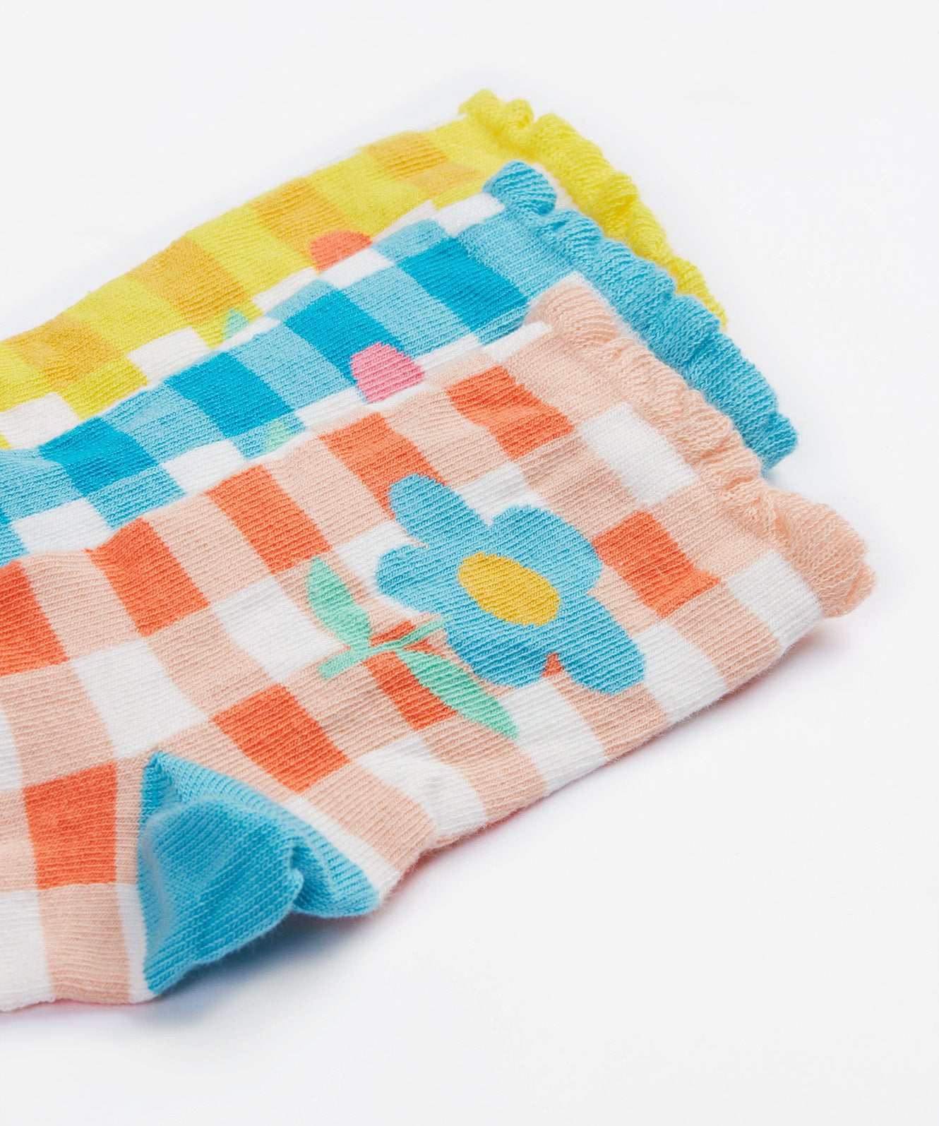 A close up of the Frugi kids Freya frilled gingahm socks - Showing the blue gingham with pink daisy sock, a yellow gingham with light orange daisy sock and a light orange gingham and blue daisy sock on organic cotton fabric