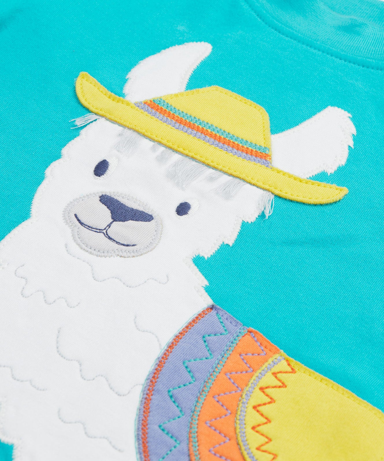 Close up of the Frugi kids easy on llama outfit. Showing the detail of the grey llama wearing a yellow hat with a purple orange and yellow saddle. Features loose cotton threads to represent hair on blue organic cotton fabric