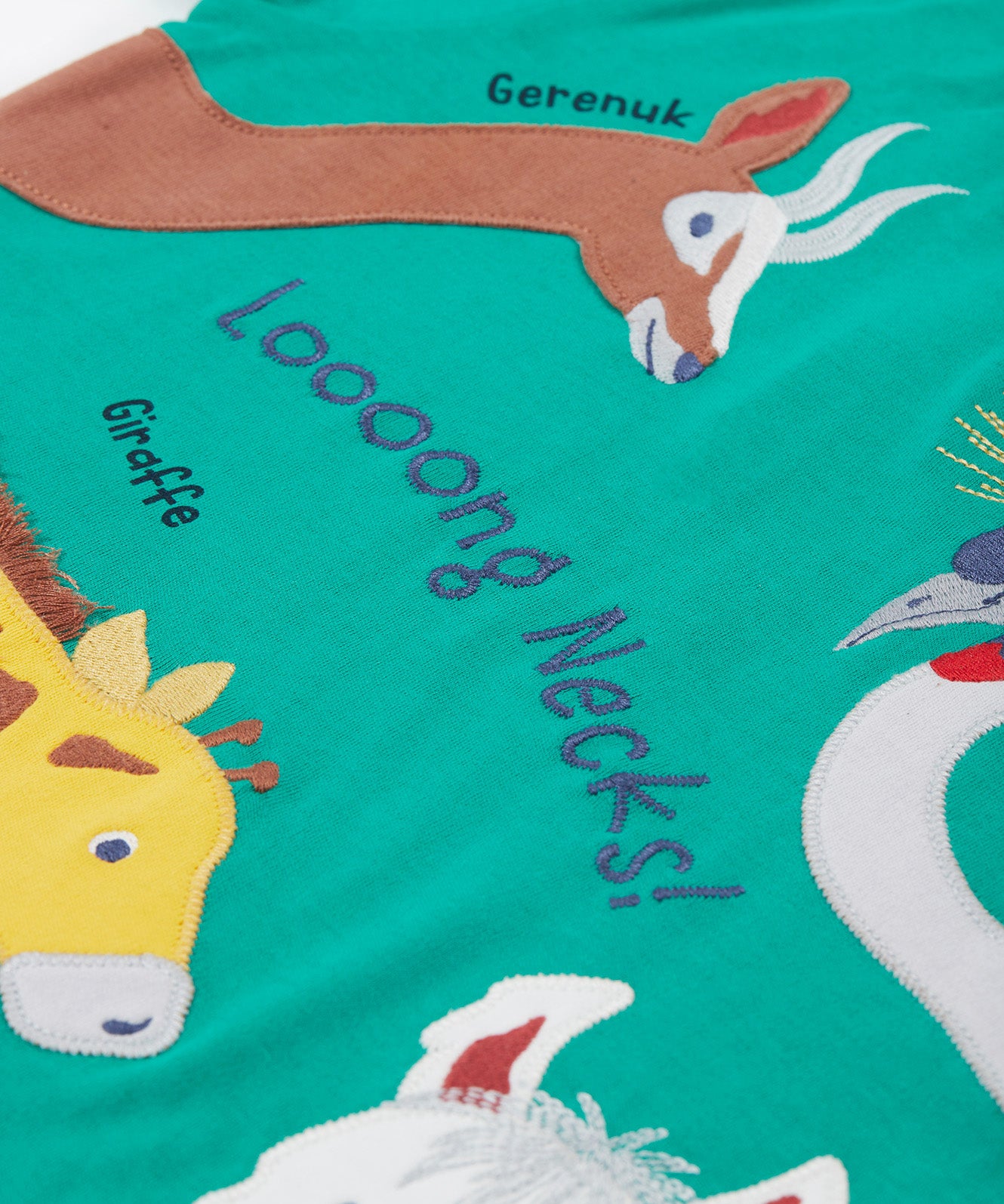 A close up of the Frugi kids long neck pals t-shirt. Showing the different long necked animals appliqué including a giraffe and gerenuk with long neck slogan across the t-shirt and animal names on green organic cotton fabric