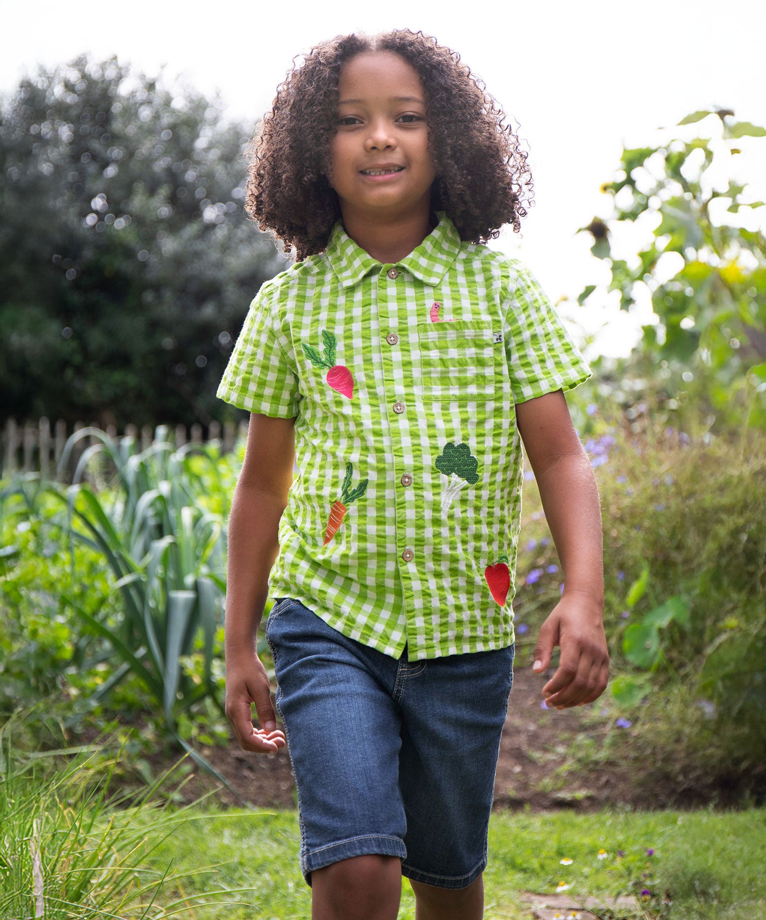 A child wearing the Frugi Green Macaw Gingham  Greengrocer Reggie Shirt with denim shorts.