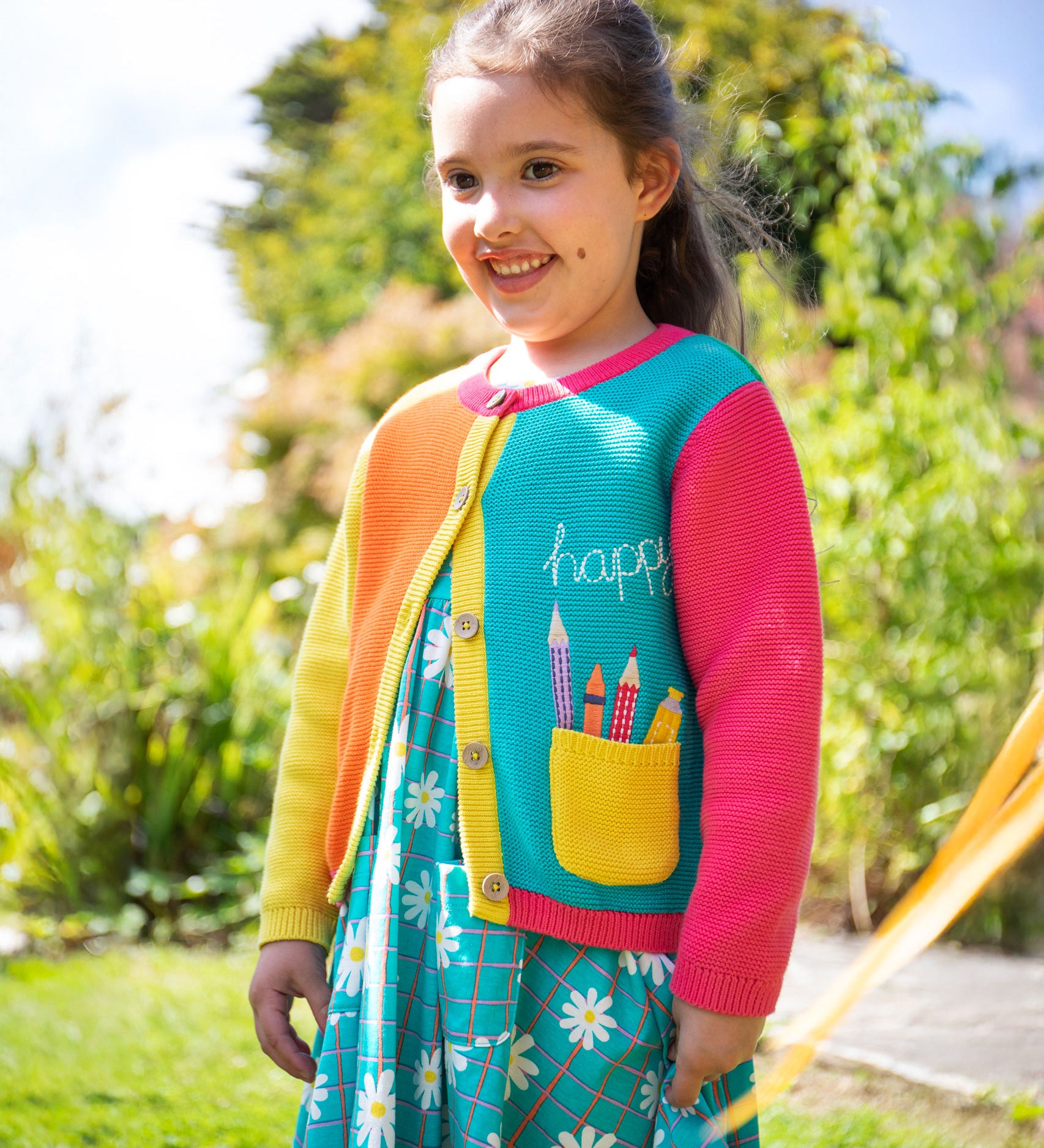 A child wearing the Frugi Happy Pen Pot Rose Pocket Cardigan with a Frugi Floral Check Summer Holiday Dress underneath.