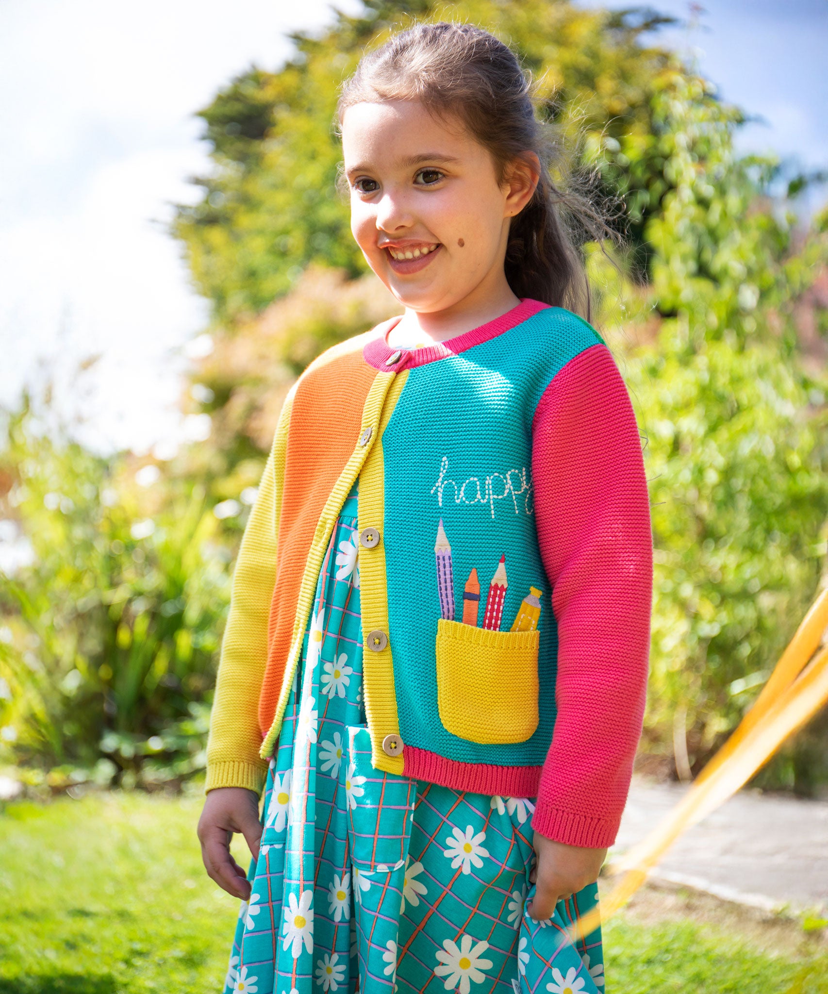 A child wearing the Frugi Happy Pen Pot Rose Pocket Cardigan with a Frugi Floral Check Summer Holiday Dress underneath.