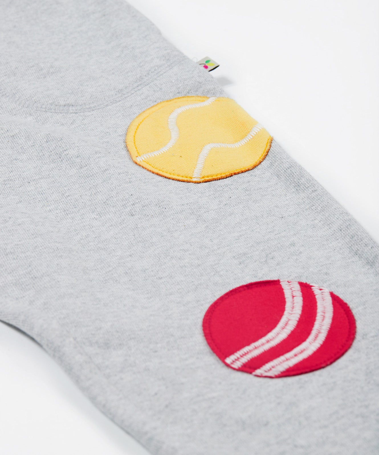 Close up of the Frugi kids jack appliqué joggers. Showing the tennis and cricket ball appliqué on grey organic cotton fabric