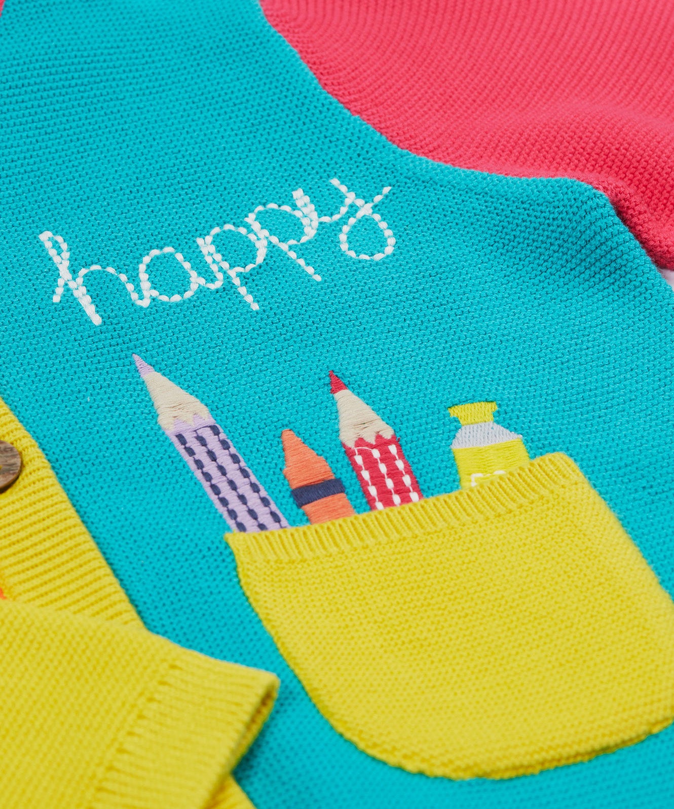 Close up of the Frugi kids Rose pocket cardigan, happy pen pot. Showing the yellow pocket with an applique of pens and pencils with a happy slogan embroidery on organic knitted cotton fabric.