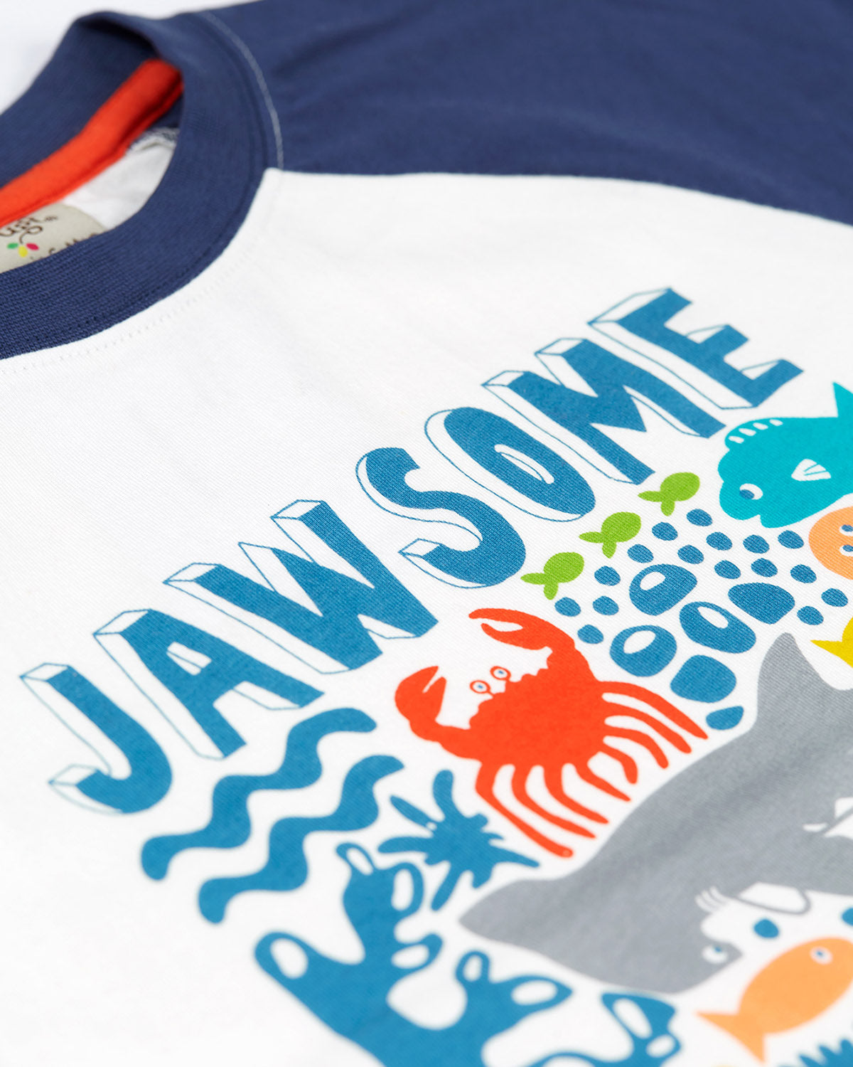 A smiling child wearing the Frugi Reid Raglan T-Shirt - Jawsome. A colourful t-shirt made from GOTS Organic cotton  with a white body with sea-life animal print