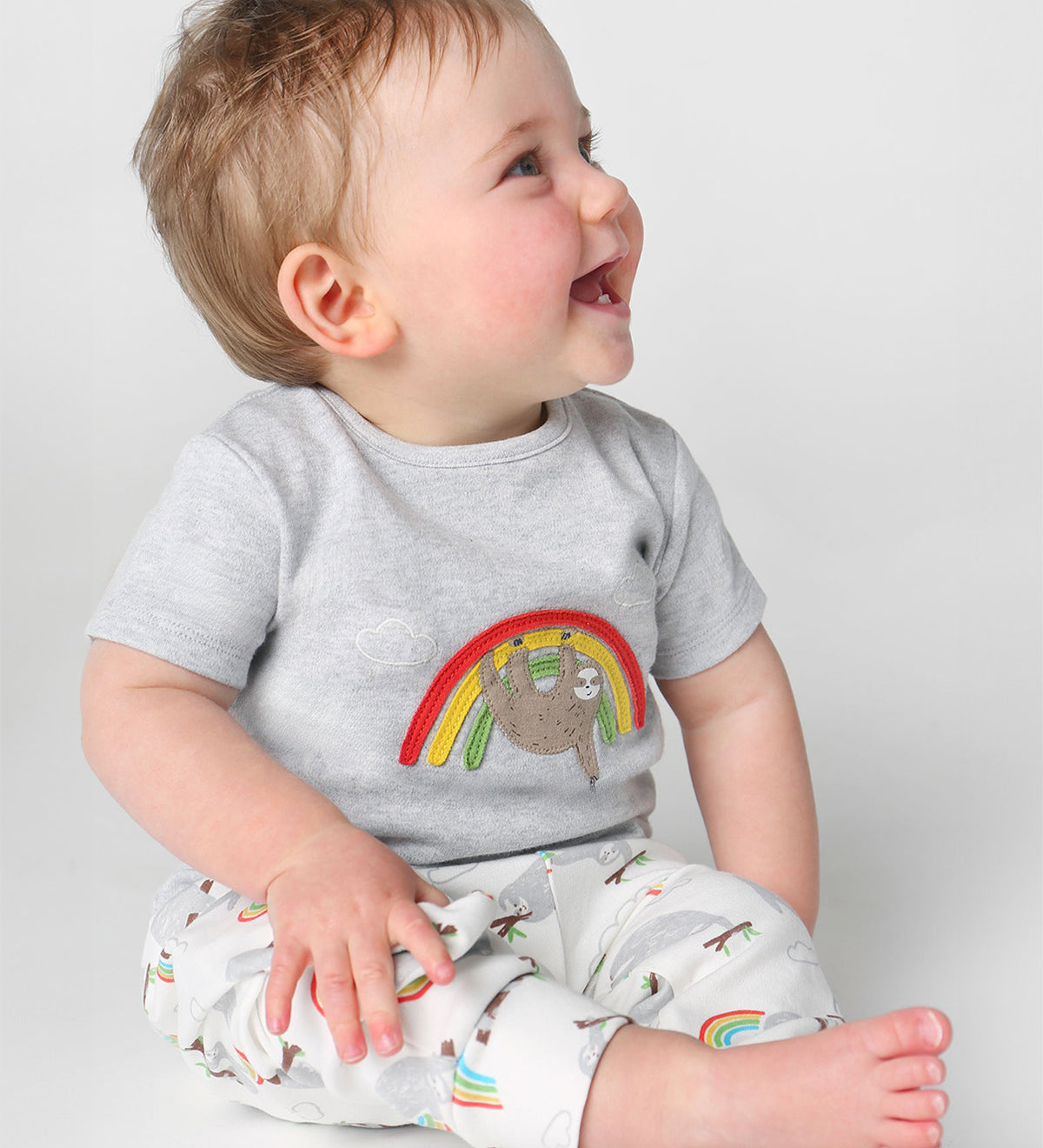 A happy toddler sat down and wearing the Frugi Children's GOTS Organic Cotton Frankie Summer Outfit - Sleepy Sloths. A beautiful 2 piece outfit including a grey short sleeved top with a sloth and rainbow character applique with comfy white cuffed sloth an