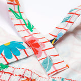 Close up of the Frugi kids Delpi paint a floral dress. Showing the back of the shoulder strap detail, coloured flowers and yellow bees on white check with red outline organic cotton fabric