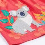 Close up of the Frugi kids flora fun koala tunic top. Showing the appliqué of a grey koala next to bamboo shoots on light pink organic cotton fabric with pastel orange floral prints