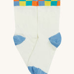 Close up of the Frugi white sunnie socks. Showing the multicoloured check design on the ankle cuff. Features a light blue heel and toe contrast panel on white organic cotton fabric