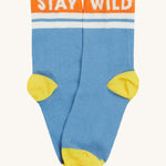 Close up of the Frugi light blue sunnie socks. Showing the 'stay wild' slogan on the orange ankle cuff. Features a light yellow heel and toe contrast panel on light blue organic cotton fabric