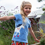 A child playing outside wearing the Frugi Elise wrap around t-shirt . A cornflower blue coloured t-shirt with with short sleeves. Features an appliqué of a white horse wrapped around the front and back of the t-shirt.