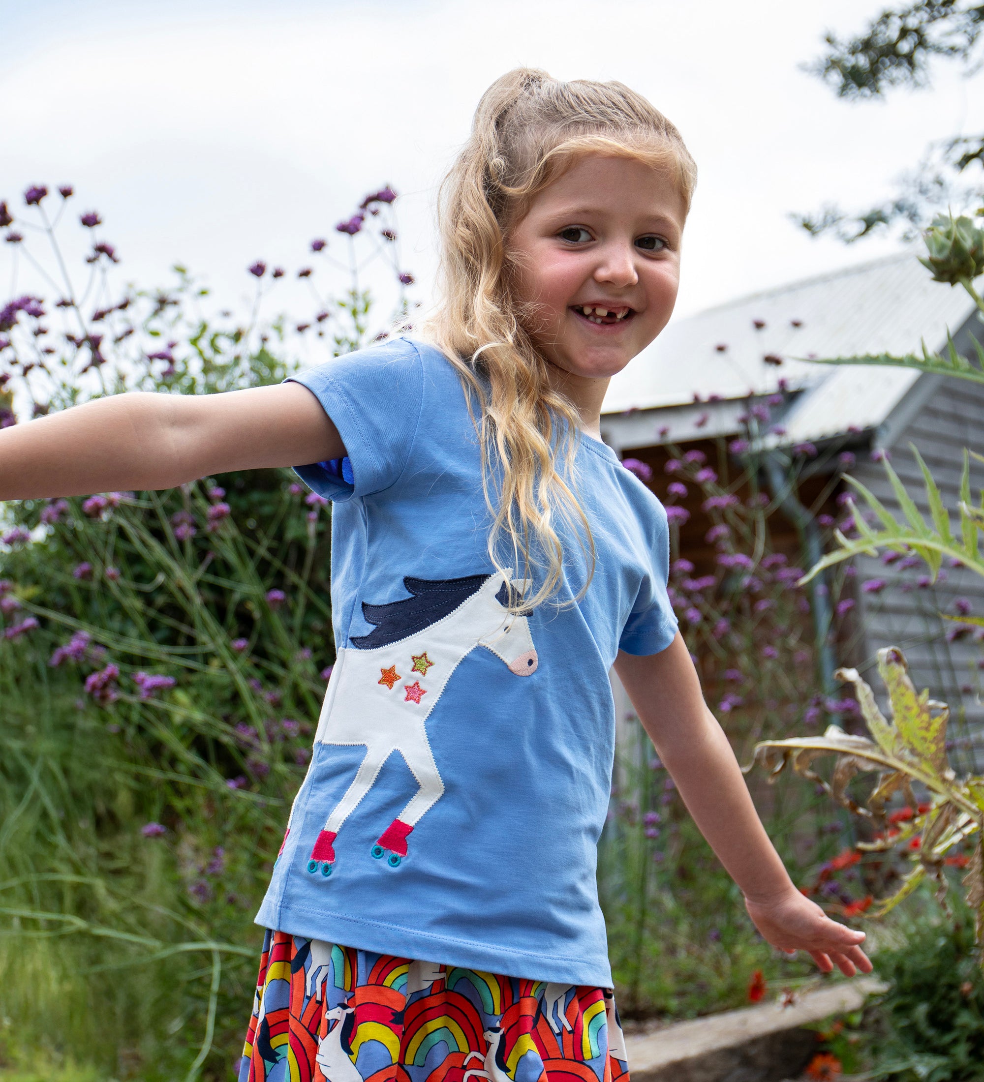 A child playing outside wearing the Frugi Elise wrap around t-shirt . A cornflower blue coloured t-shirt with with short sleeves. Features an appliqué of a white horse wrapped around the front and back of the t-shirt.