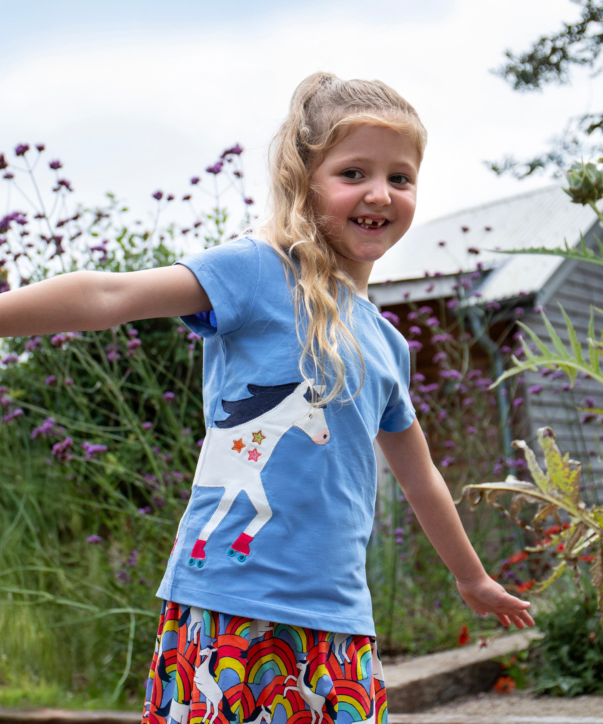 A child playing outside wearing the Frugi Elise wrap around t-shirt . A cornflower blue coloured t-shirt with with short sleeves. Features an appliqué of a white horse wrapped around the front and back of the t-shirt.