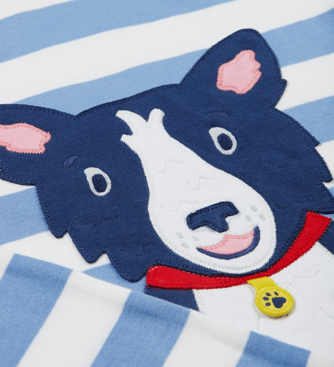 A close up of the Frugi kids Porth collie dog pyjama set showing the appliqué of a collie dog on blue and white striped organic cotton fabric