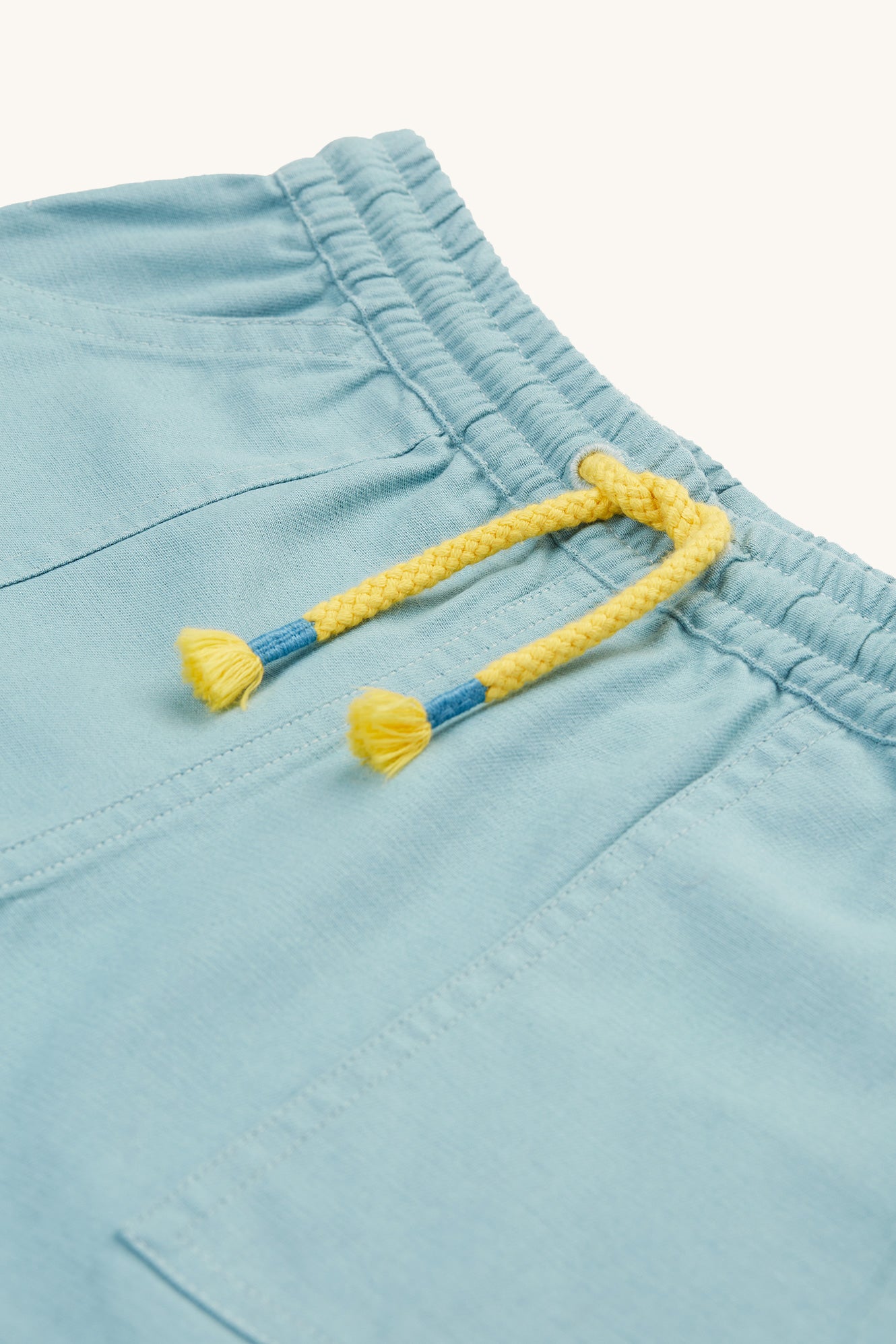 A closer look of the Plain Blue side of the Frugi Children Organic Cotton Rocky Reversible Shorts - Stingray / Jawsome. A plain pale blue short with yellow drawstring on one side and a Jawsom shark outline print on the reverse