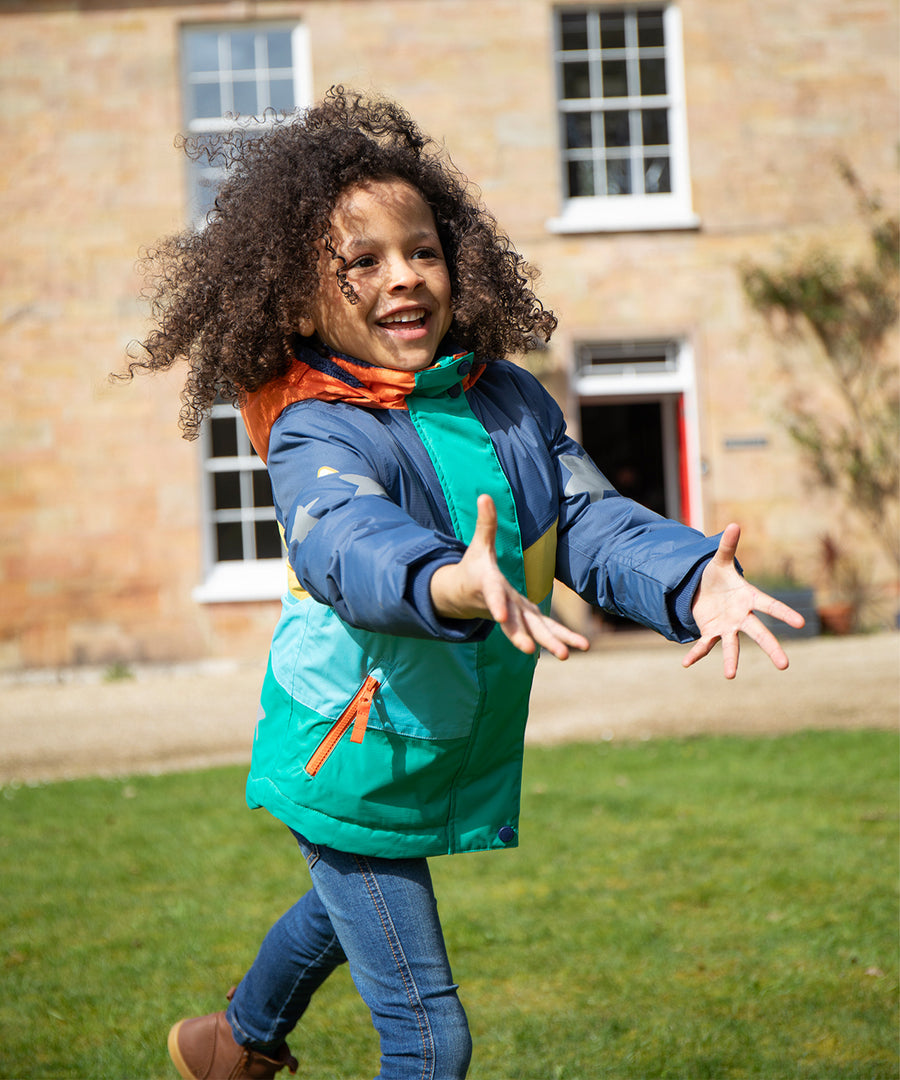 A child happily outside wearing the Frugi Snow & Ski Coat - Iguana Colourblock. The image shows the zipper at the front open