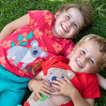 Children lying on the grass. One child wearing the Frugi Carsen t-shirt - a red with short sleeves and a playful appliqué of a brown quokka. Frugi Lottie t-shirt - a watermelon pink with short sleeve and an appliqué of a grey koala 