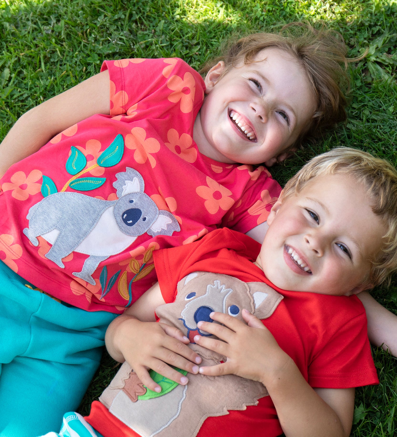 Children lying on the grass. One child wearing the Frugi Carsen t-shirt - a red with short sleeves and a playful appliqué of a brown quokka. Frugi Lottie t-shirt - a watermelon pink with short sleeve and an appliqué of a grey koala 