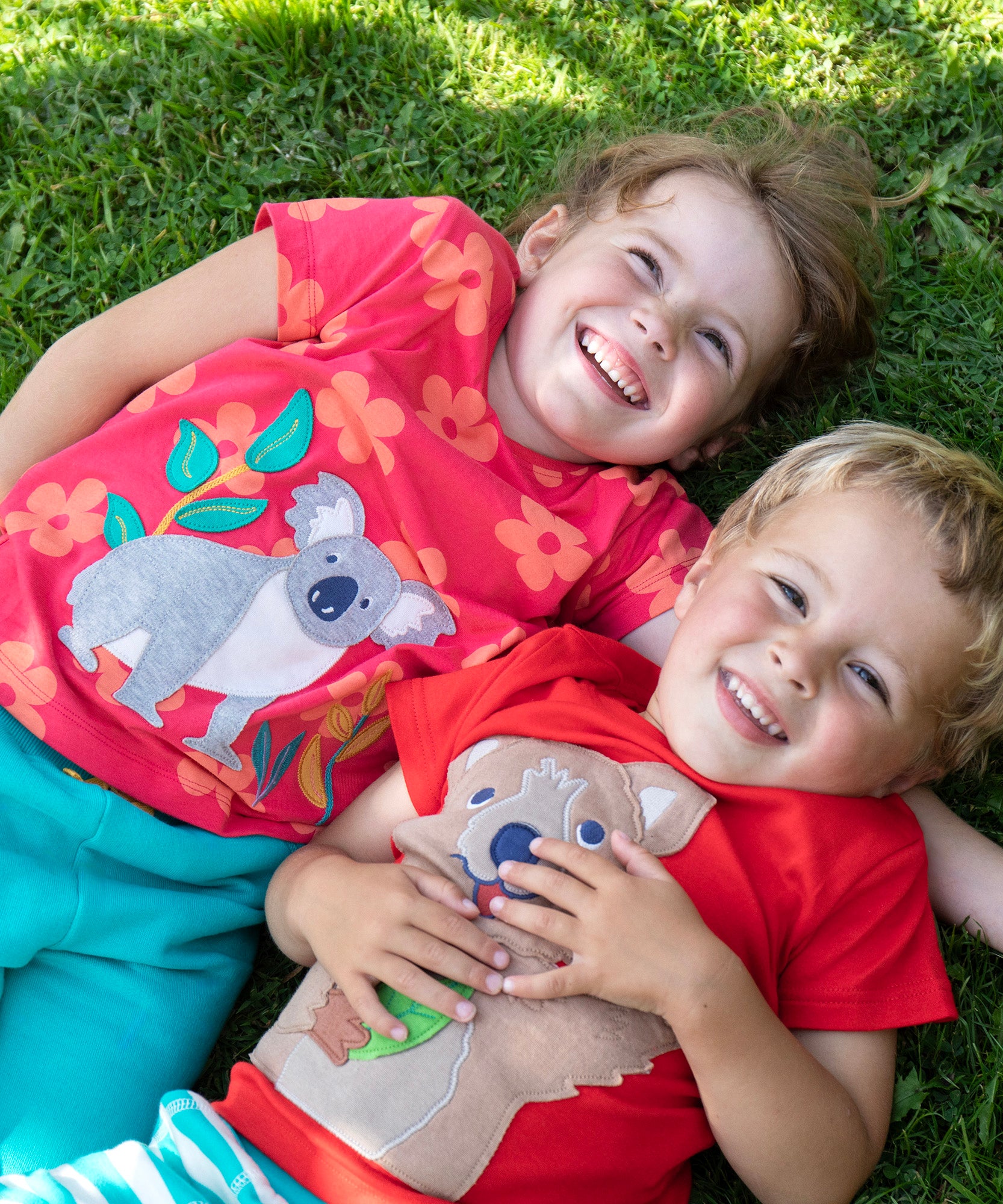 Children lying on the grass. One child wearing the Frugi Carsen t-shirt - a red with short sleeves and a playful appliqué of a brown quokka. Frugi Lottie t-shirt - a watermelon pink with short sleeve and an appliqué of a grey koala 