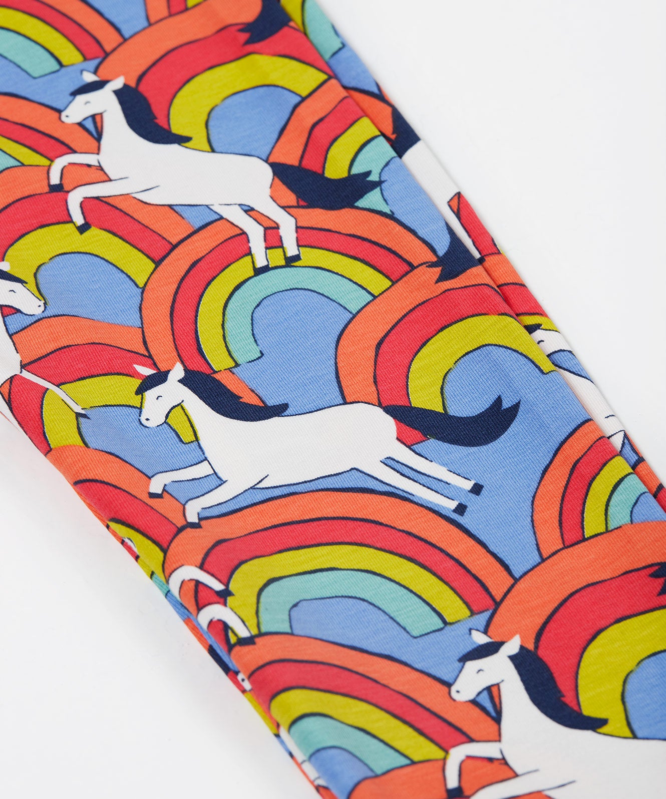 Close up of the Frugi kids over the rainbow leggings. Showing the white horses on top of bright multicoloured rainbows on light blue organic cotton fabric