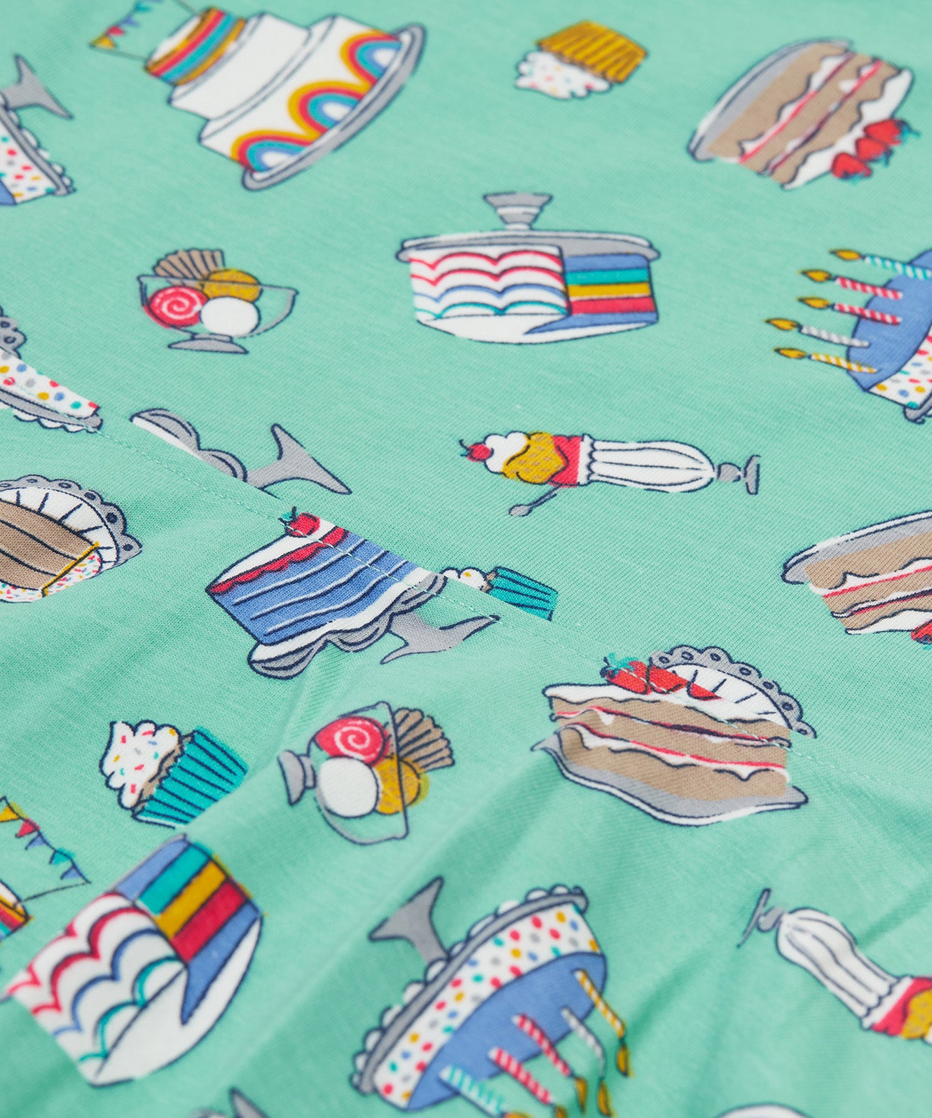 A close up of the Frugi kids lets bake Morwenna skater dress. Showing the different cakes, ice cream, puddings on light green organic cotton fabric