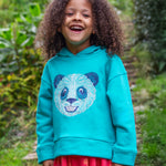 A child standing outside wearing the Frugi oversized Lissie hoody - a light blue colour with a playful panda embroidery on the front. They are wearing the Frugi Samantha Floral Fun / Flamingo Summer Dress underneath the hoody.