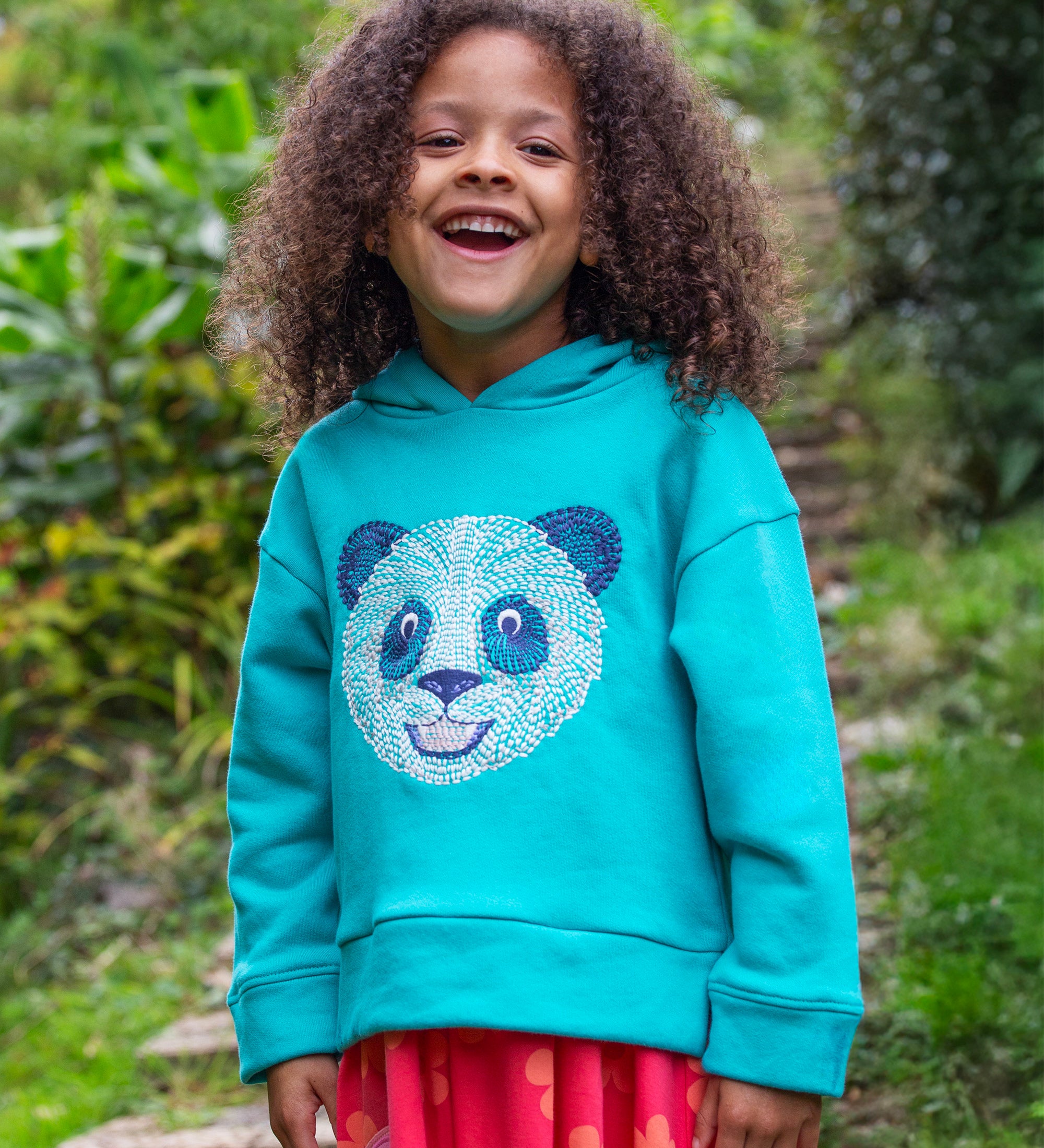 A child standing outside wearing the Frugi oversized Lissie hoody - a light blue colour with a playful panda embroidery on the front. They are wearing the Frugi Samantha Floral Fun / Flamingo Summer Dress underneath the hoody.