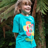 A child wearing the Frugi  Happy Days Mabe Relaxed T-Shirt. A turquoise blue organic cotton t-shirt with a large flower applique design on the front.The words Happy Days are placed on the centre of the flower applique. 