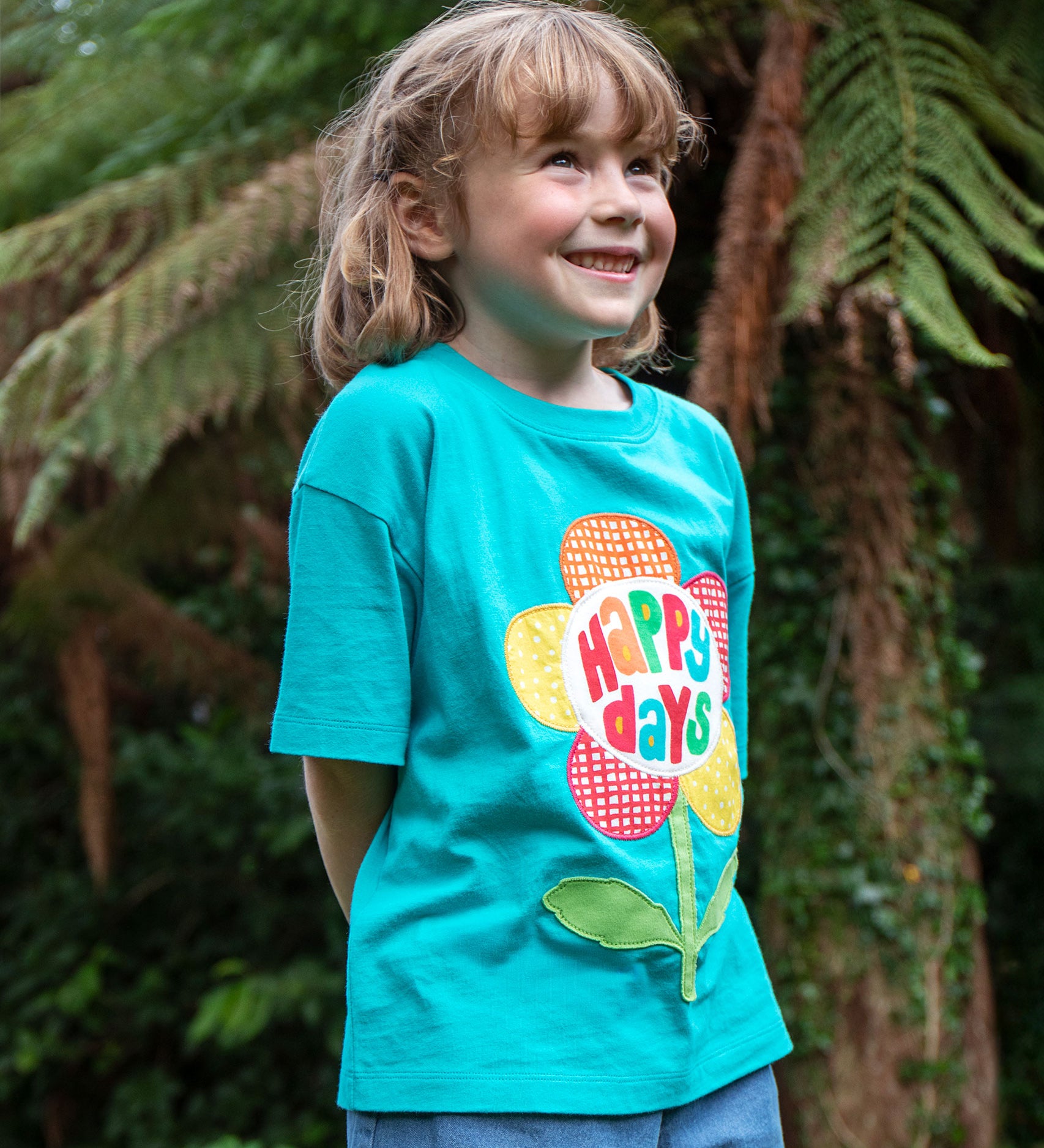 A child wearing the Frugi  Happy Days Mabe Relaxed T-Shirt. A turquoise blue organic cotton t-shirt with a large flower applique design on the front.The words Happy Days are placed on the centre of the flower applique. 