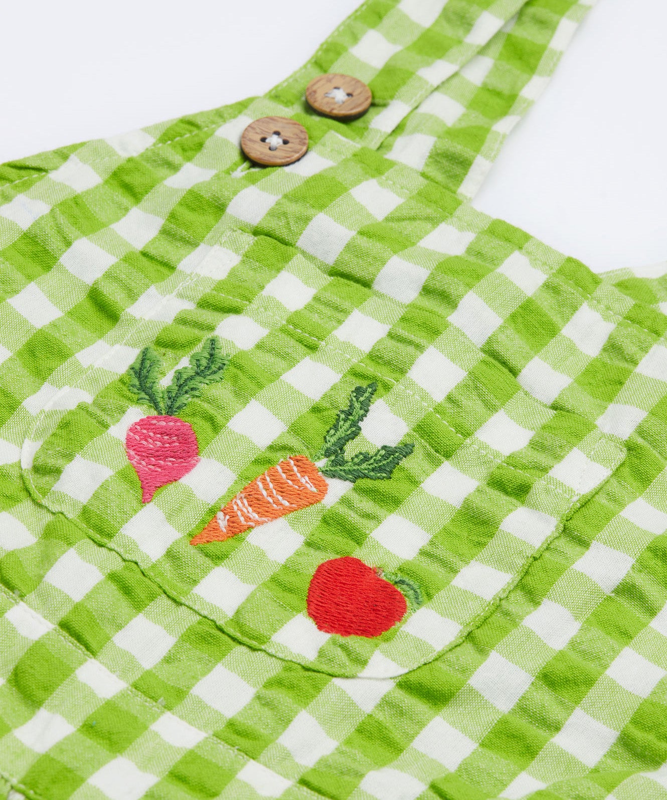 Close up of the Frugi kids macaw ginham, veg culotte dungarees. Showing the embroidery of a beetroot, carrot and apple on the front pouch pocket on green and white gingham organic cotton fabric