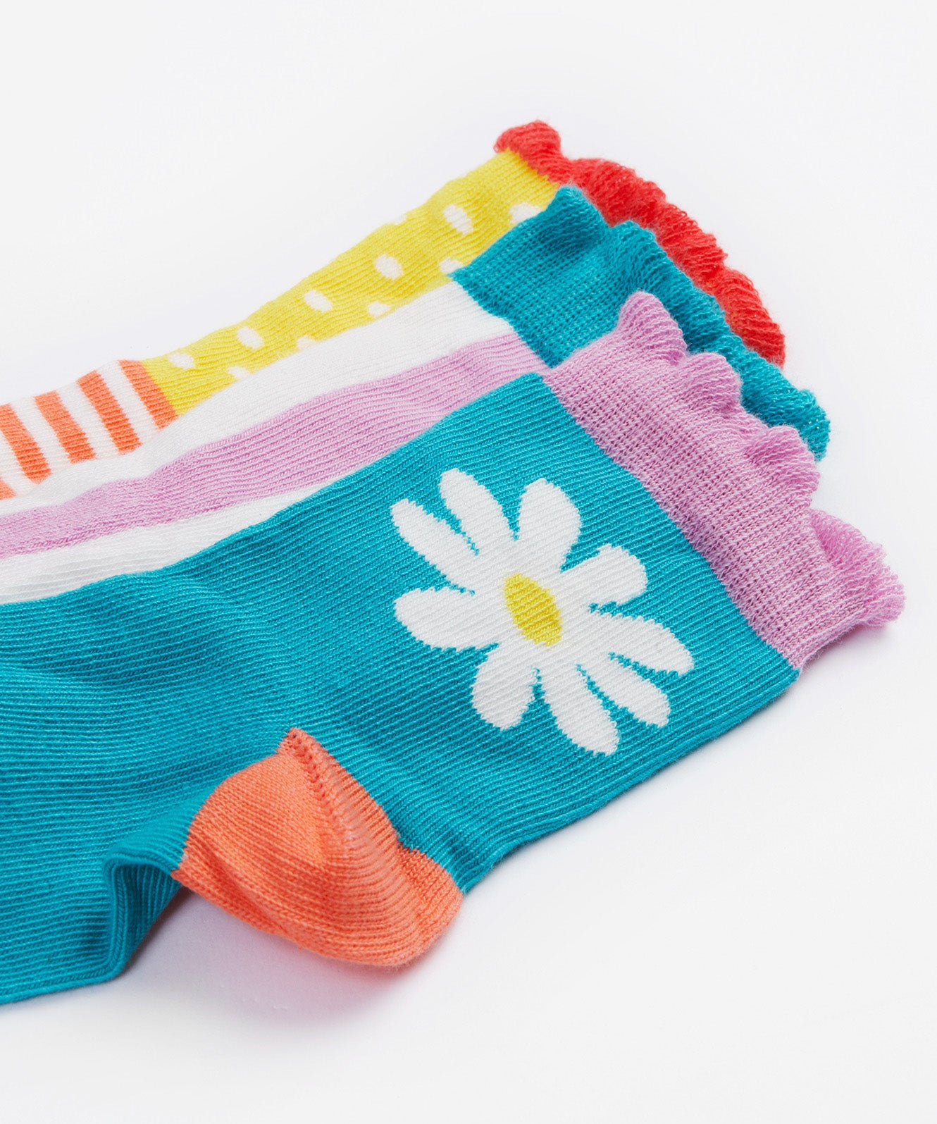 A close up of the Frugi kids Freya frilled let's create 3 pack frilled socks - Showing the  striped socks with a light blue cuff, a light blue sock and white daisy design and a spotted/striped sock on organic cotton fabric