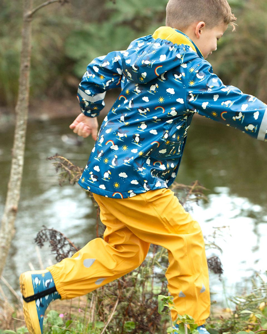 Child running in front of some water wearing the Frugi waterproof puddle buster puffin jacket and wellies