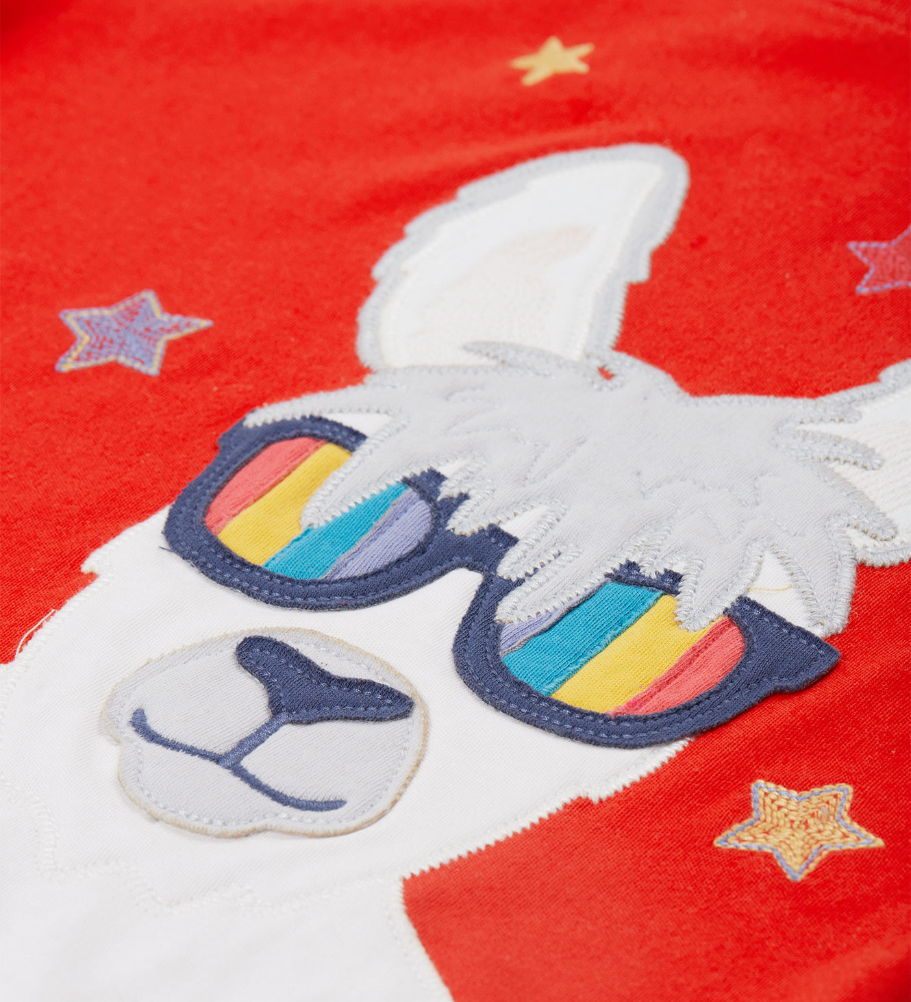 Close up of the Frugi kids red llama character t-shirt showing the white and grey llama appliqué wearing rainbow sunglasses and surrounded by different coloured embroidered stars on red organic cotton fabric