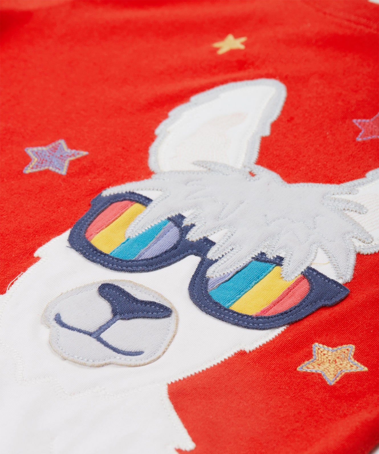 Close up of the Frugi kids red llama character t-shirt showing the white and grey llama appliqué wearing rainbow sunglasses and surrounded by different coloured embroidered stars on red organic cotton fabric