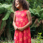 A child standing on grass outside wearing the Frugi Samantha summer dress - a light pink colour with a orange floral print and a flamingo appliqué 